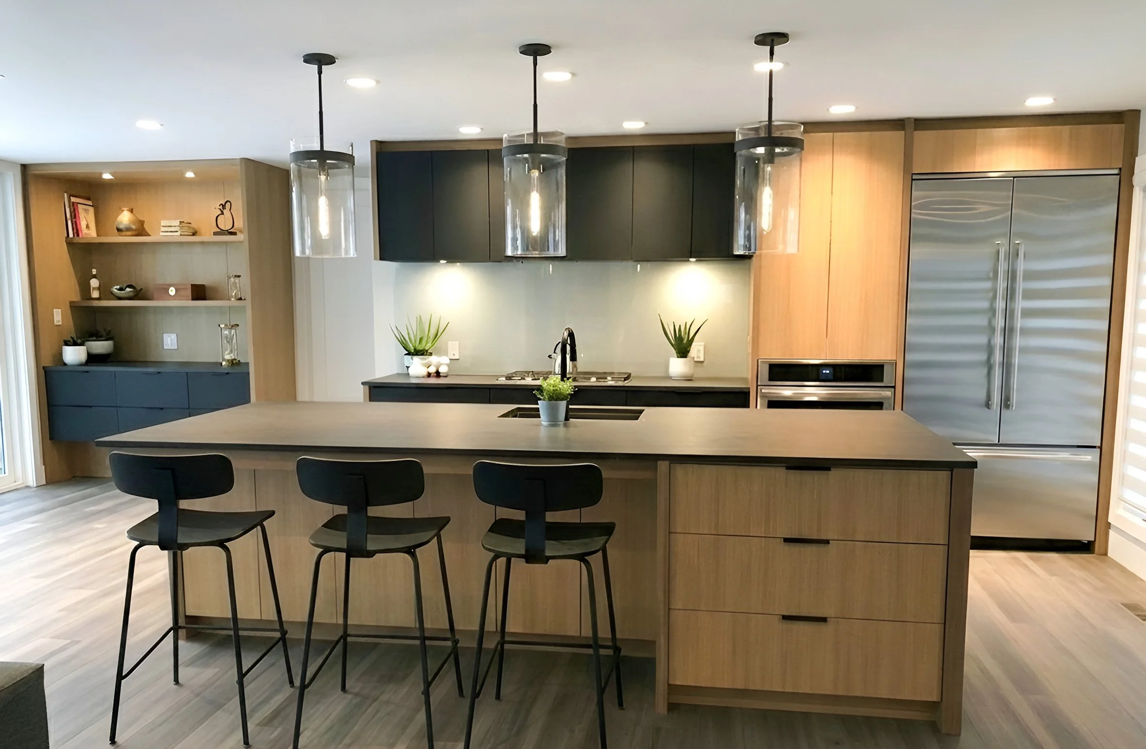 London, Ontario Main floor kitchen renovation Scandi Modern kitchen with wooden cabinetry, black upper cabinets, stainless steel appliances, a kitchen island with black countertop, three black barstools, pendant lights, and decorative plants.