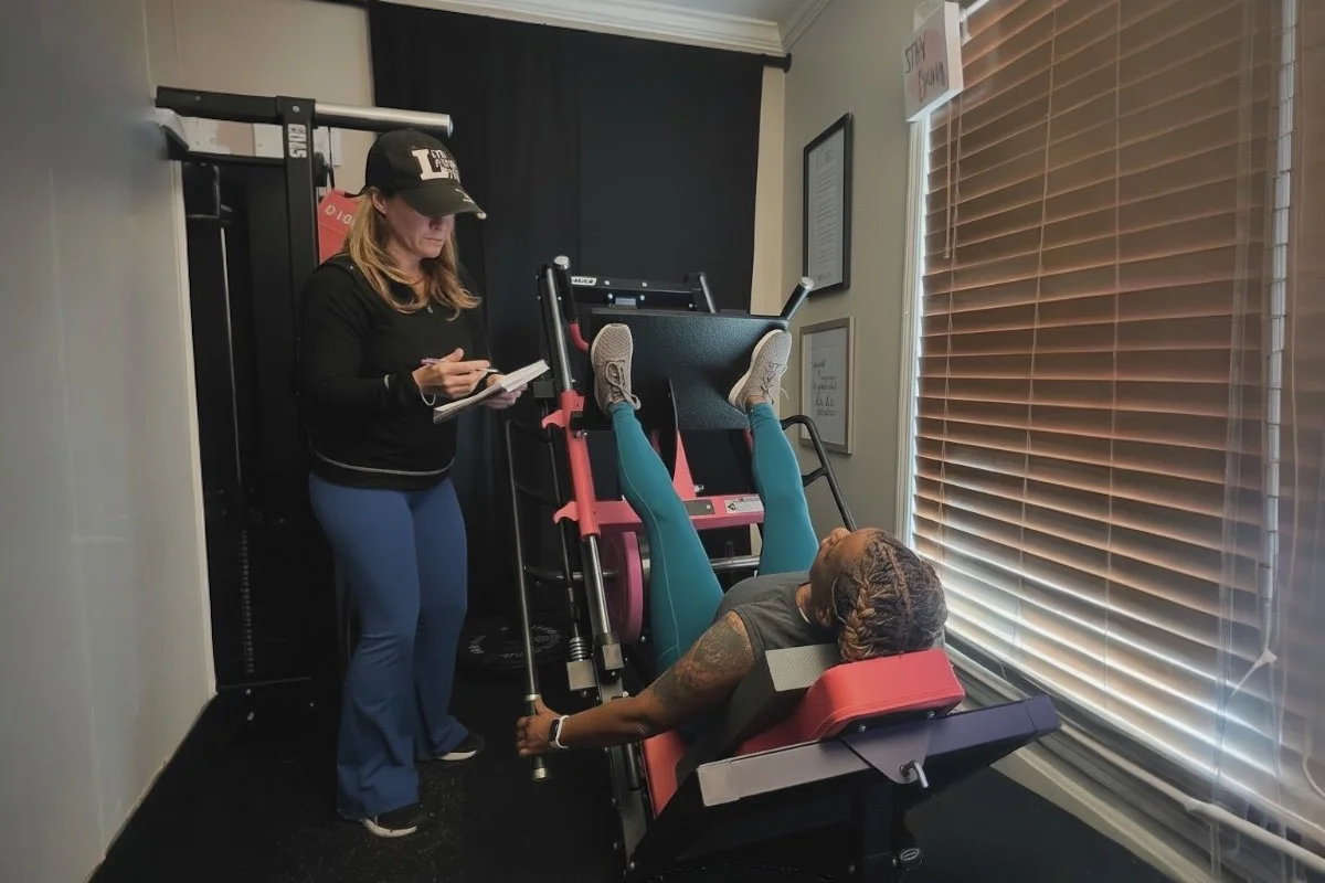 A woman exercises on a leg press machine while a trainer or staff member writes notes in a notepad nearby.