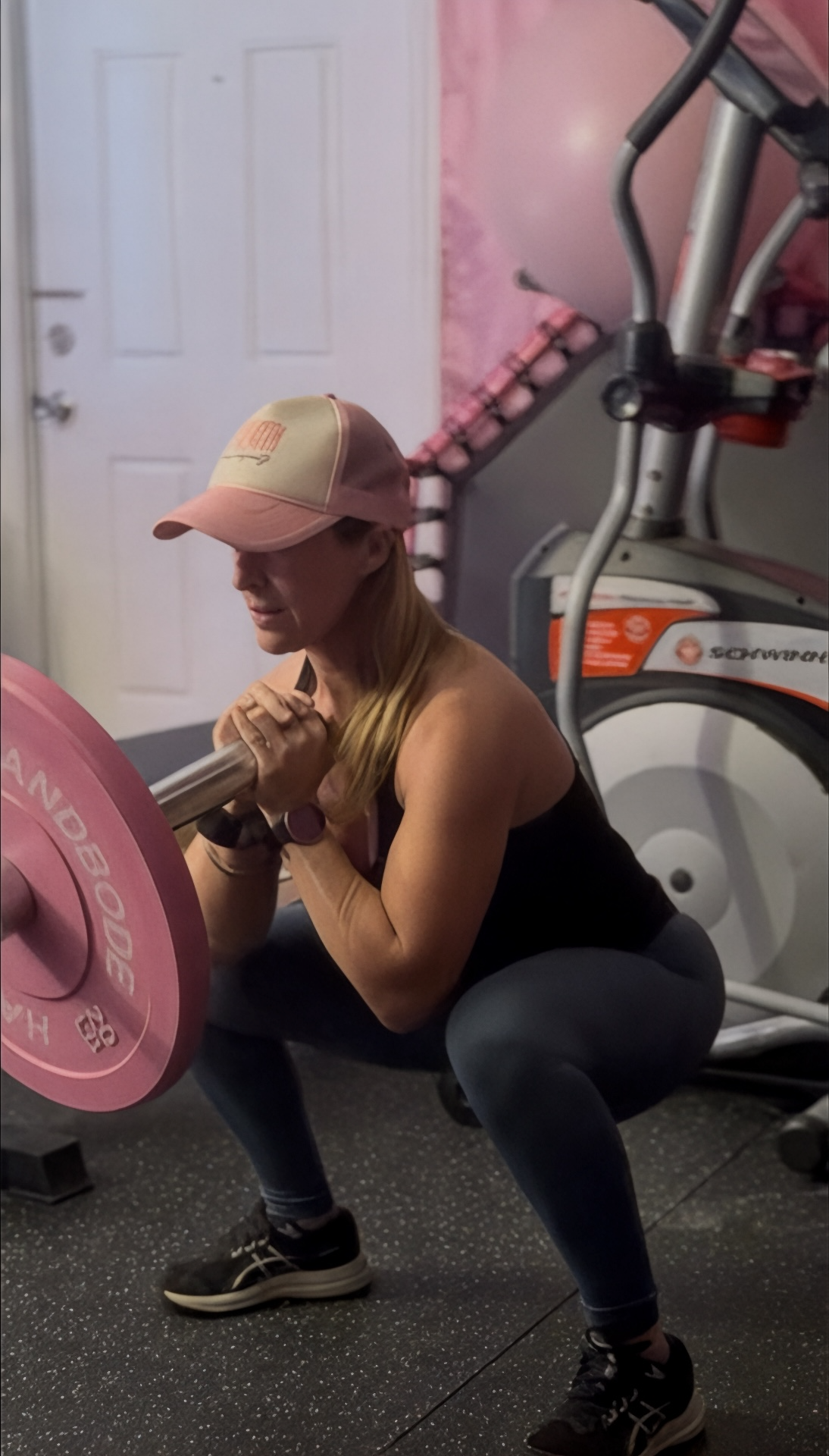 A woman in workout clothes and a pink cap is doing a squat with a barbell in a gym.