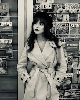 A woman with dark hair and makeup standing in front of magazine racks in black and white.