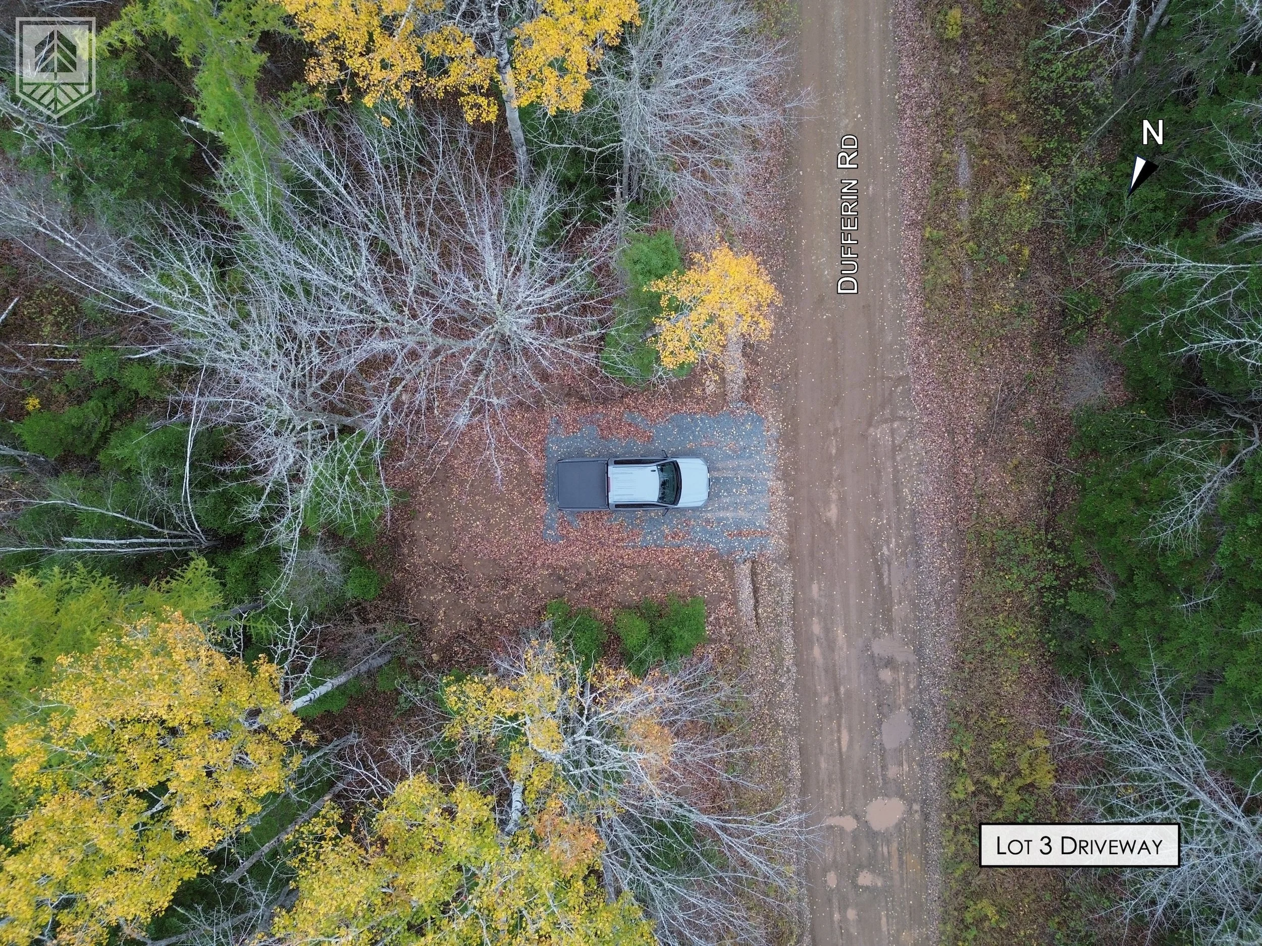 An aerial view of a dirt driveway labeled 'Lot 3 Driveway' leading to a gravel parking area with a single van parked on it. The driveway runs beside a wooded area with trees, some with yellow leaves and others bare, indicating fall.