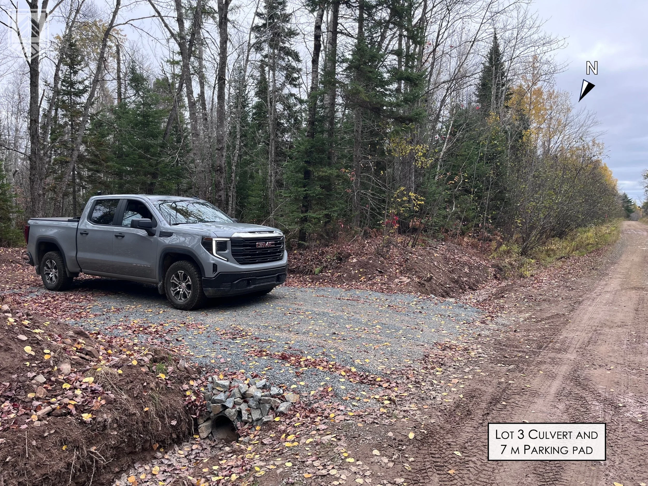A gray GMC pickup truck parked in a gravel lot with a wooded background and a dirt road on the right, surrounded by fall leaves.