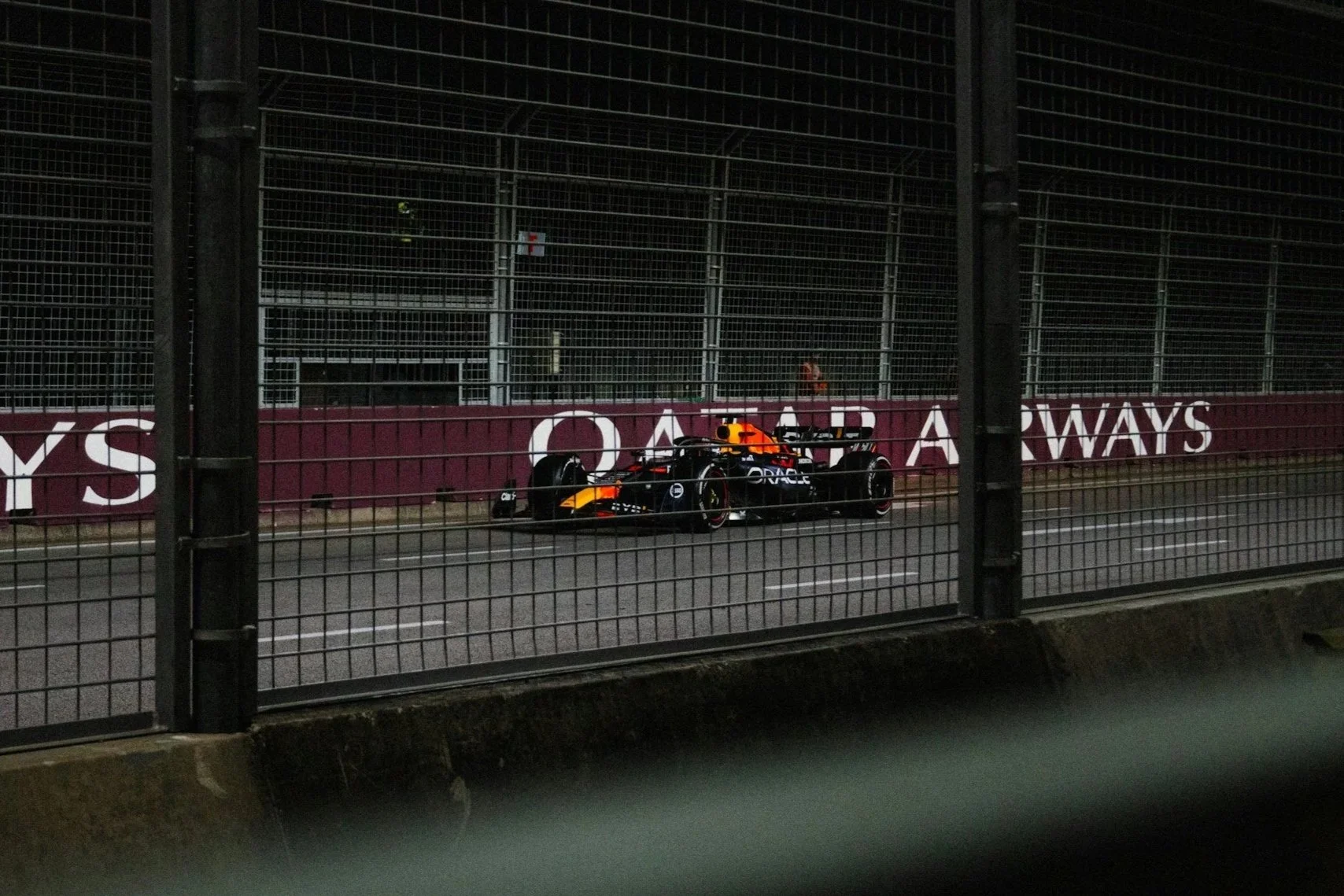 A Formula 1 race car on a dark race track, enclosed by a tall, metal safety fence.