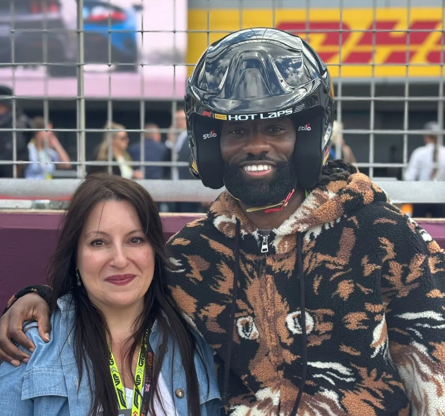 A woman with dark hair and a smiling man wearing a tiger-print hoodie and a motorcycle helmet at a racing event, smiling and posing for a photo behind a metal barrier.