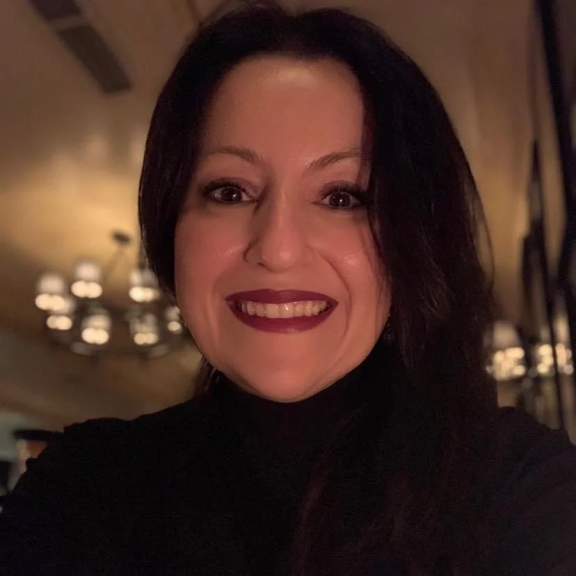 A woman with dark hair and makeup smiling in a dimly lit room with chandeliers in the background.