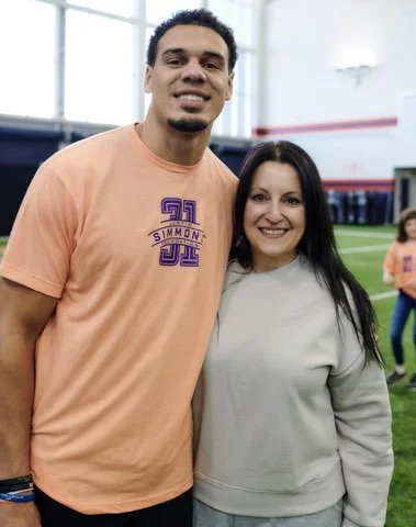 A man and woman smiling and standing close together indoors, with other people in the background and a sports field.