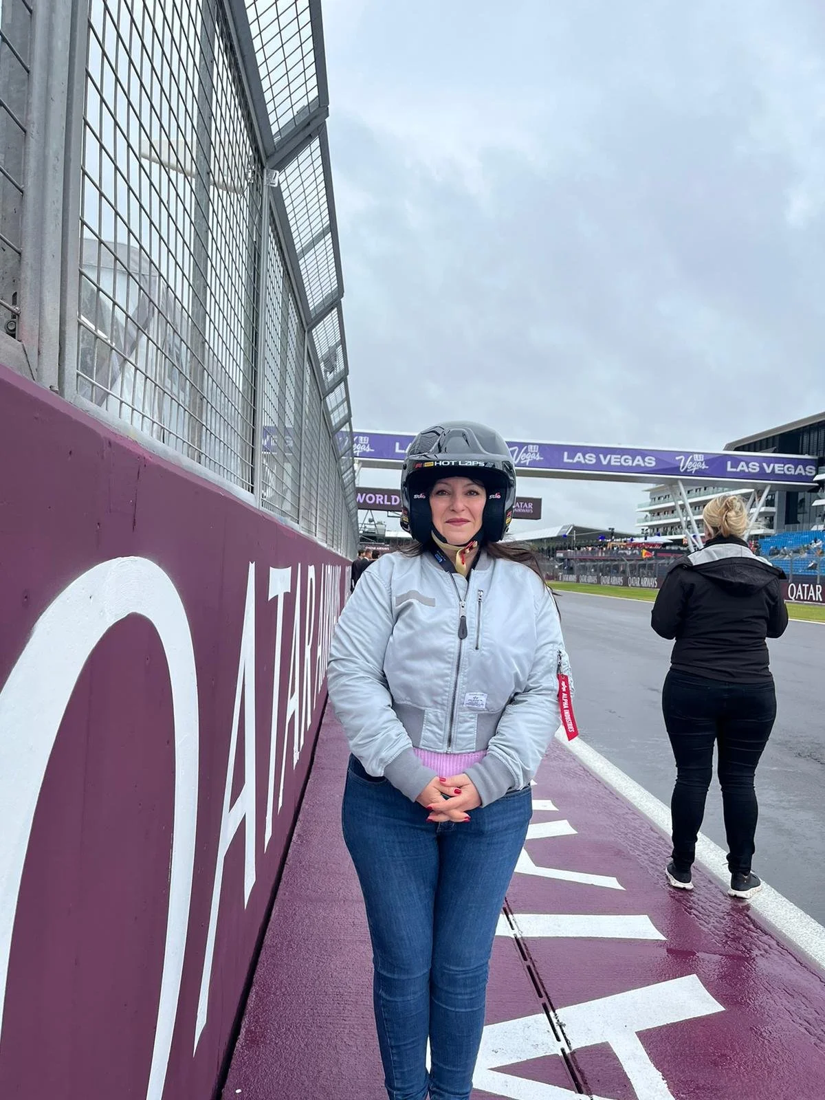 Woman wearing a helmet and a white jacket standing on a race track at Las Vegas Grand Prix, with another woman in black jacket in background and empty grandstands.