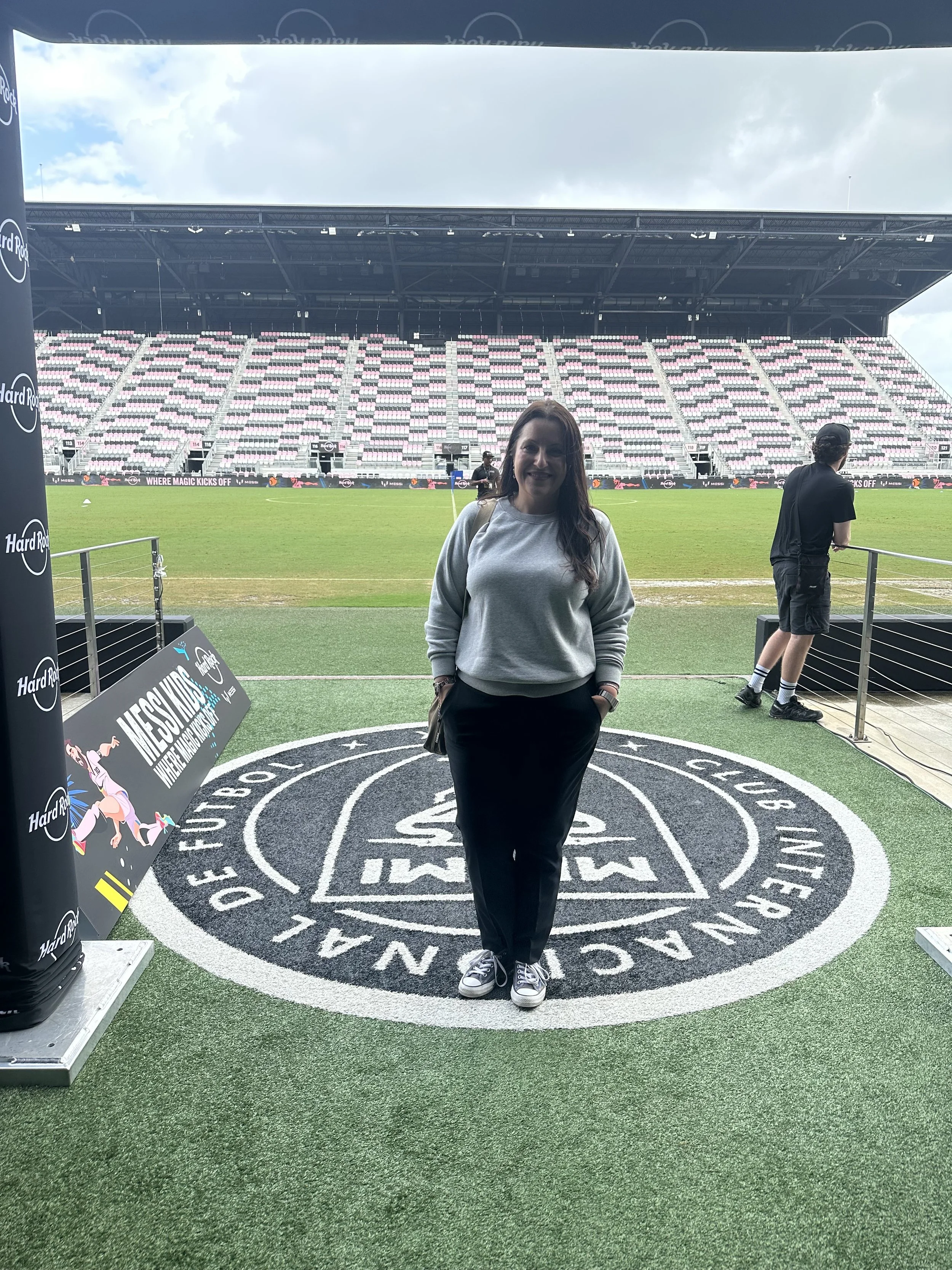 Woman standing on a circular logo on a sports field with stadium seating in the background, sunny sky with clouds, and a person standing near the railing.