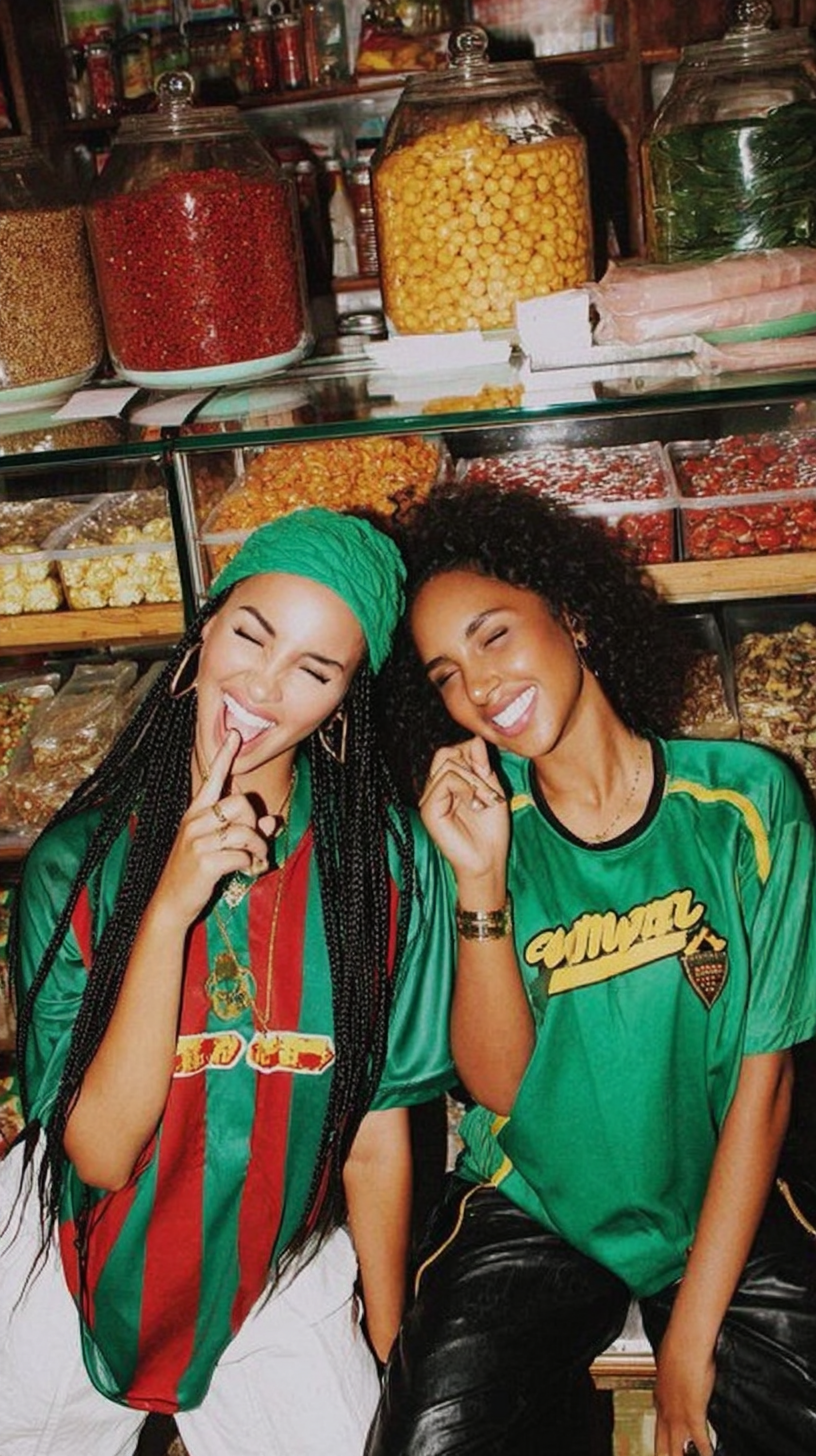 Two women smiling and posing inside a candy store, with large jars of candy on shelves behind them. One woman has braided hair and is wearing a green headwrap and colorful shirt, while the other has curly hair and is wearing a green sports jersey.