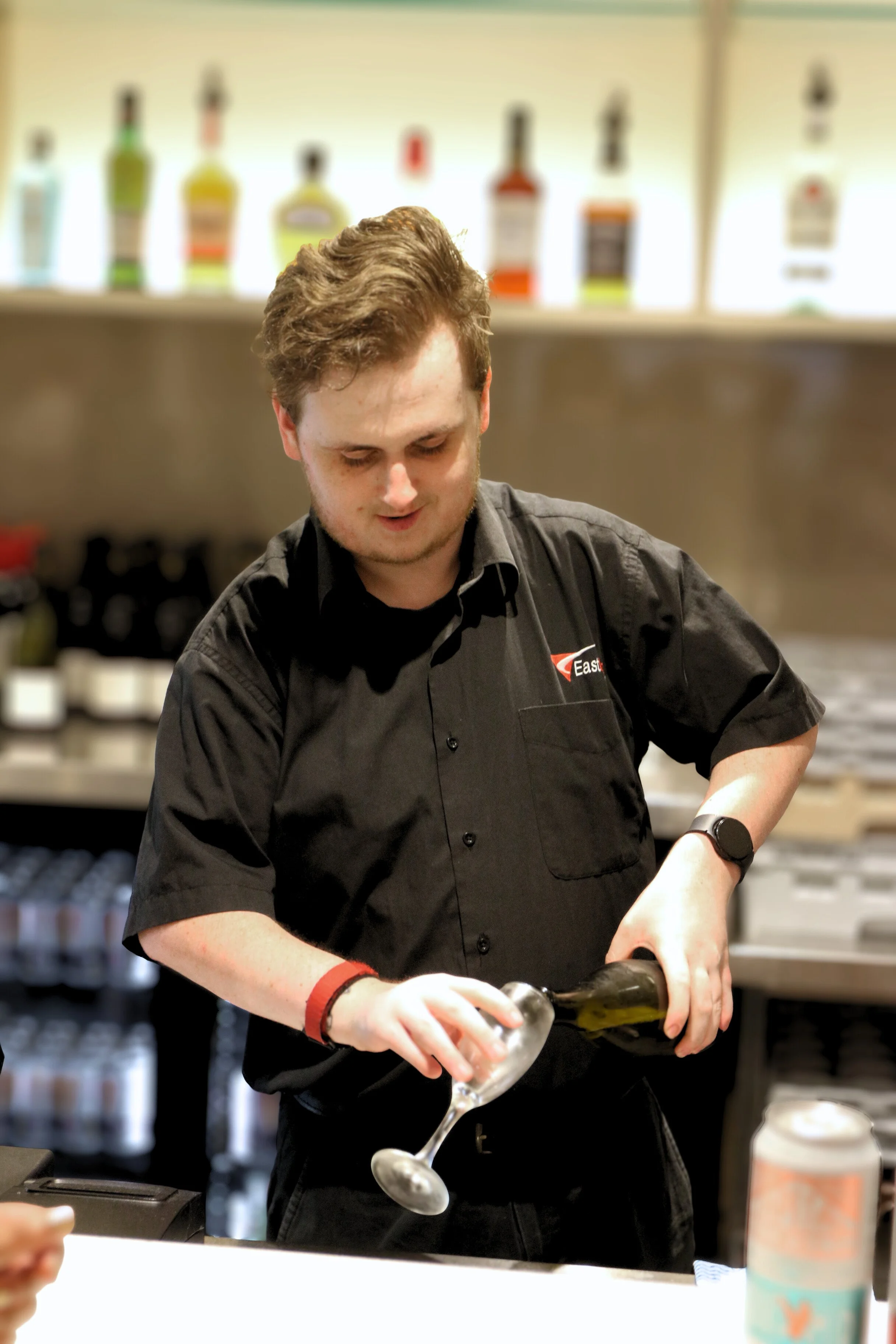 A young man in a black shirt is pouring wine into a glass in a bar or restaurant setting.