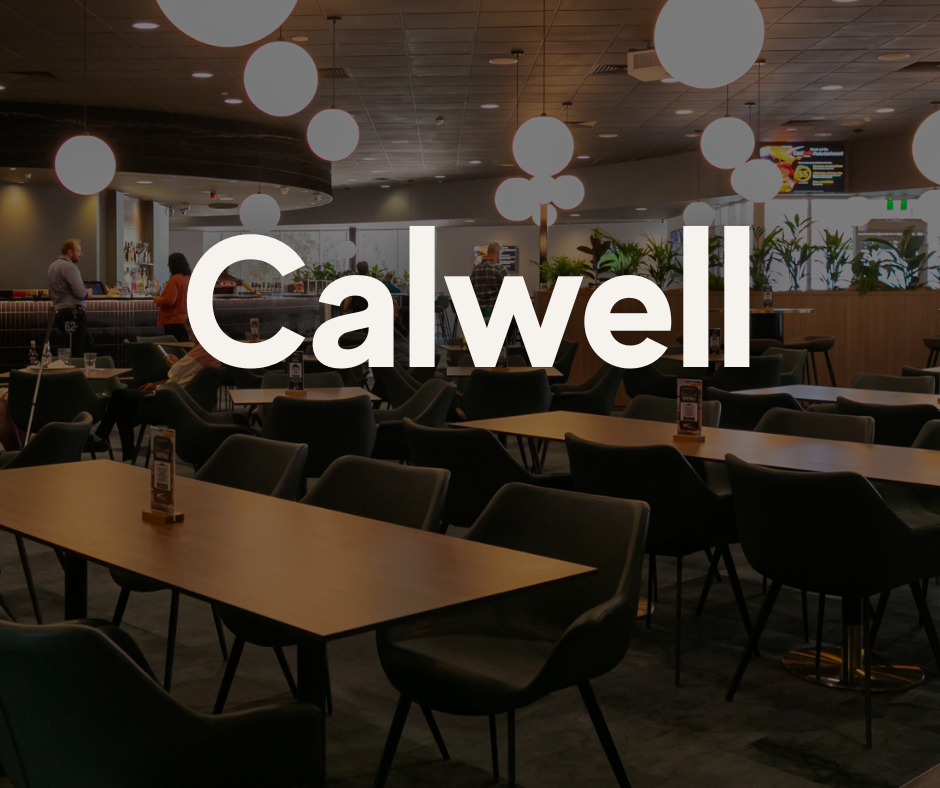 Interior of a modern restaurant or cafe with empty tables and chairs, decorative hanging lights, a bar area, and a few people standing and talking.