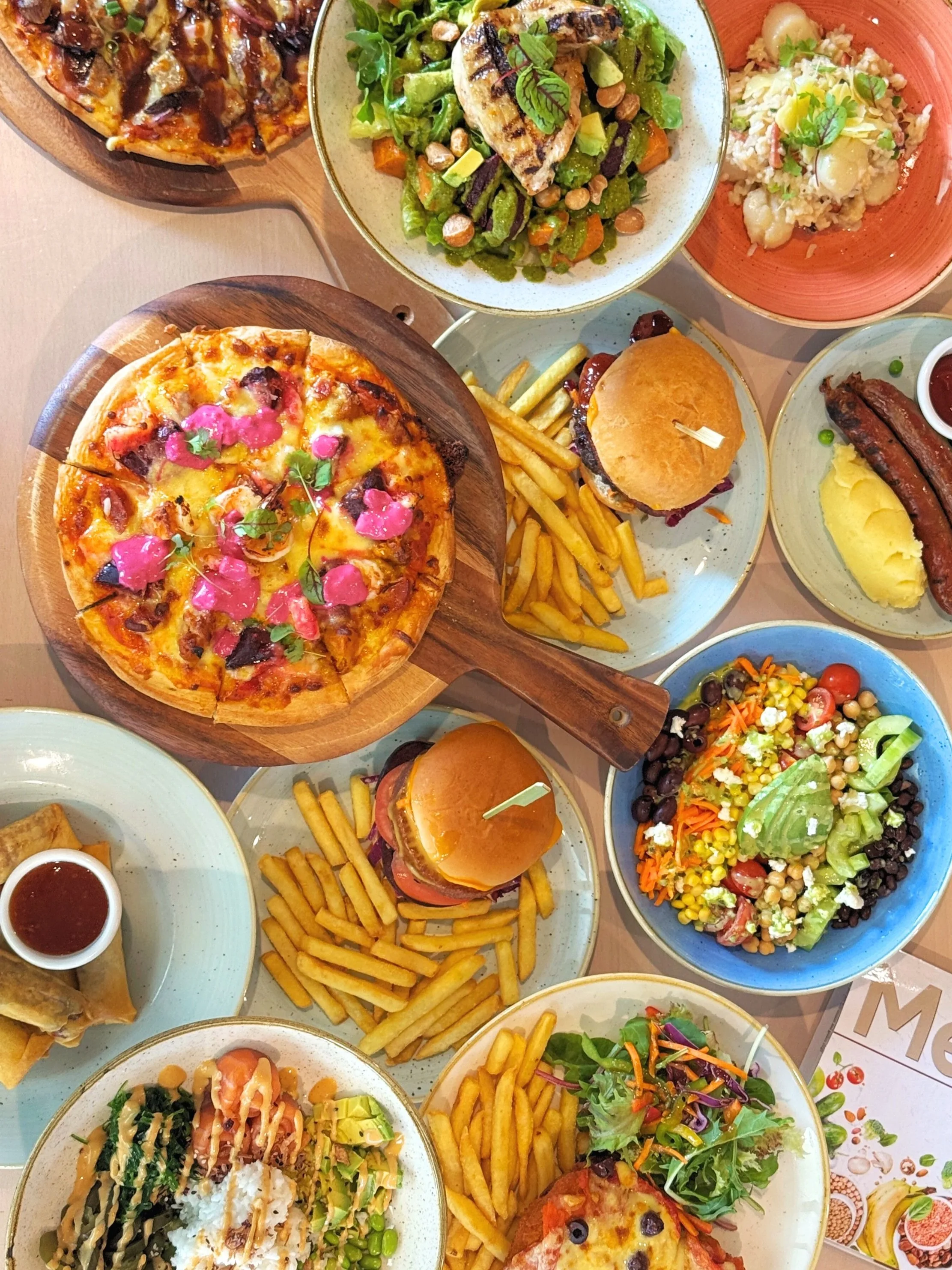 A variety of dishes including pizza, burgers, salads, potato fries, grilled sausages, and spring rolls on different plates and bowls.
