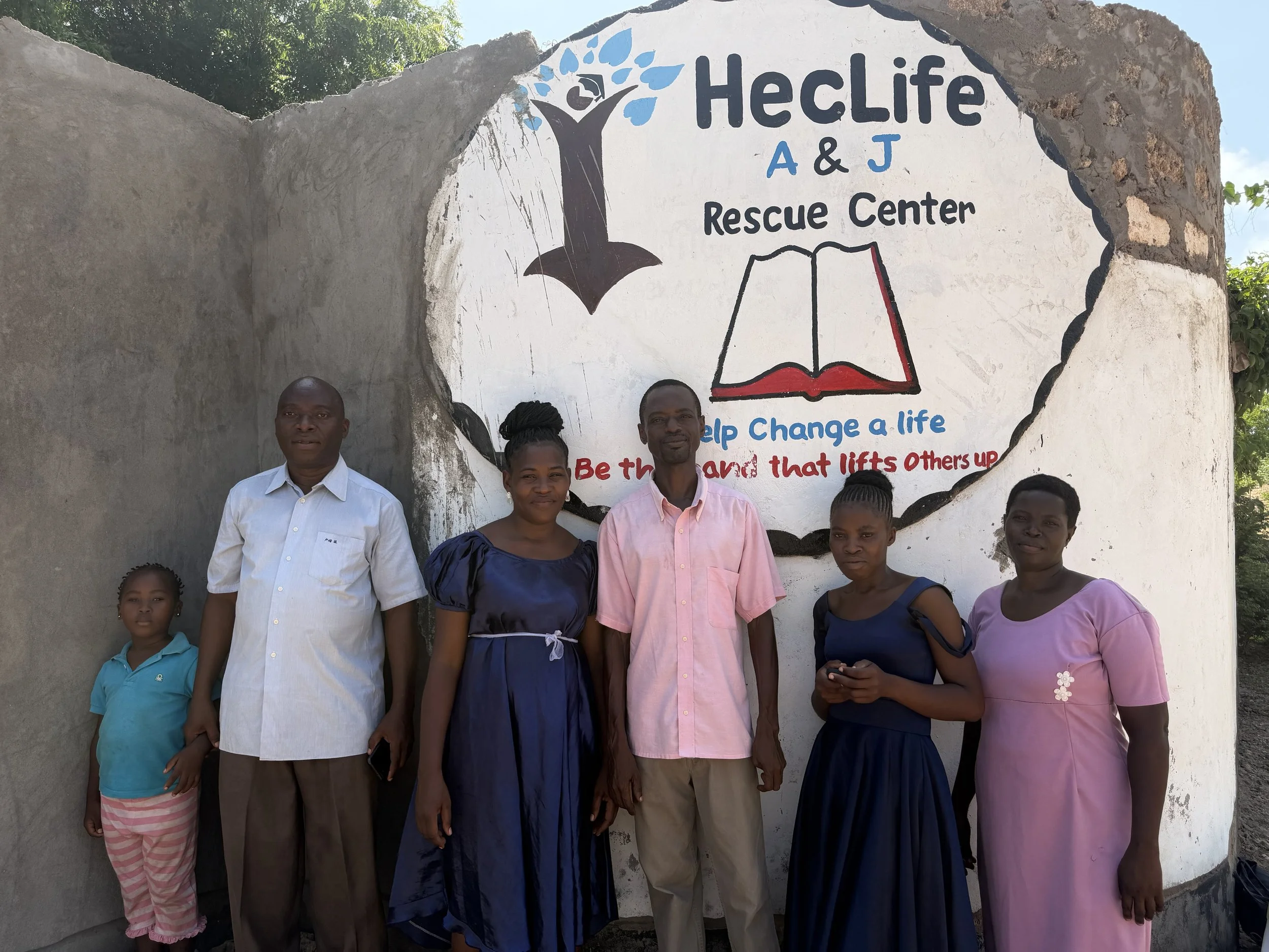 A group of six people standing in front of a mural at the HeCLife A & J Rescue Center. The mural features a tree, an open book, and a quote encouraging helping others.