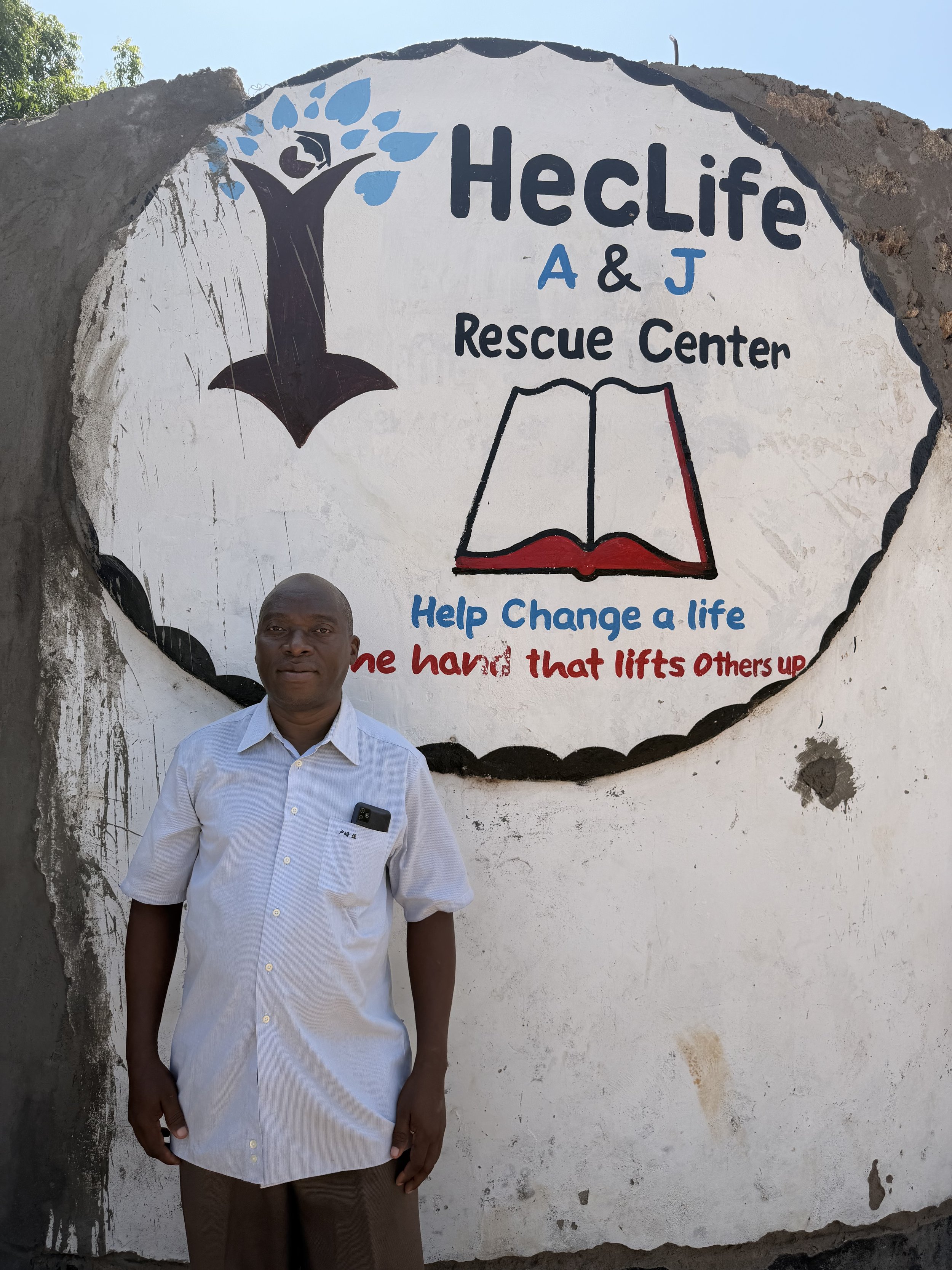 A man standing in front of a sign for Heclife A & J Rescue Center, which features a tree, an open book, and the slogan 'Help Change a life one hand that lifts others up.'