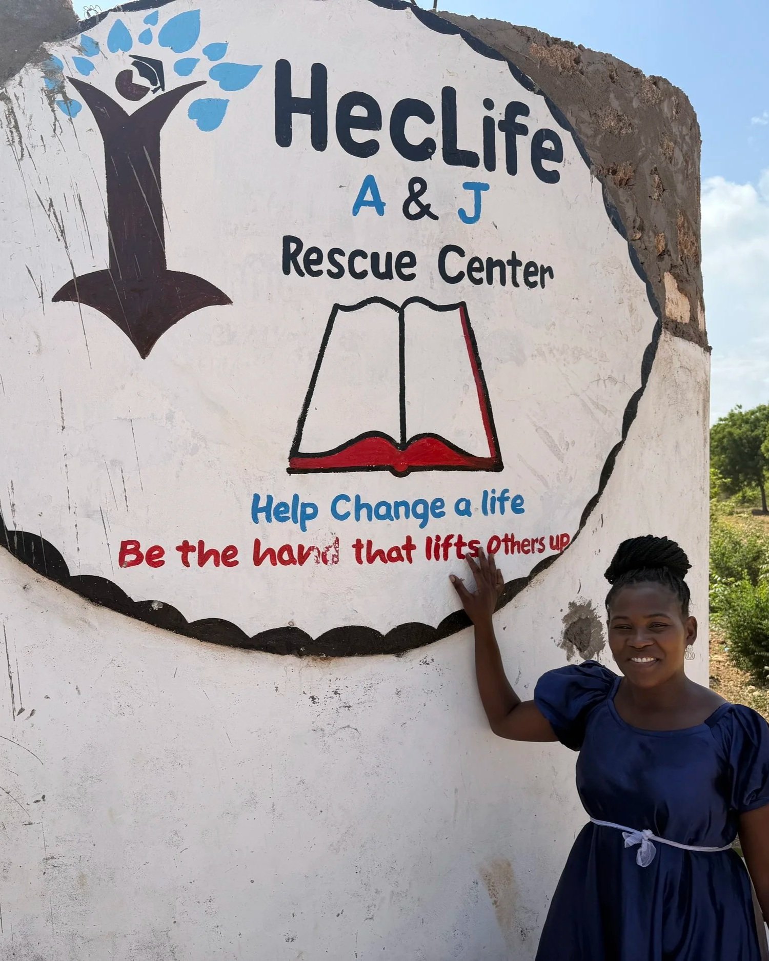 A young woman standing next to a painted sign for HeCLife A & J Rescue Center. The sign includes a tree and an open book with red edges, with the words 'Help Change a life' and 'Be the hand that lifts Others up' written on it.