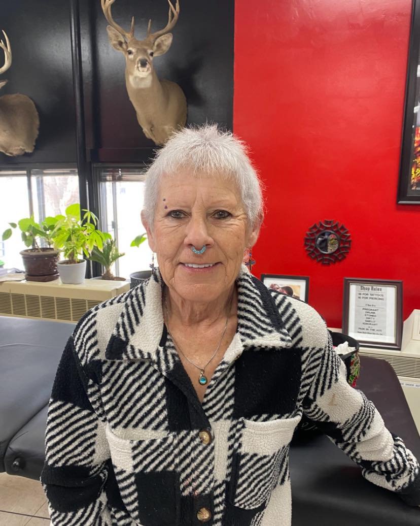 An elderly woman with short gray hair, wearing a black and white patterned jacket and turquoise jewelry, standing indoors in front of a red wall with framed photos and a mounted deer head. There are green plants and large windows behind her.