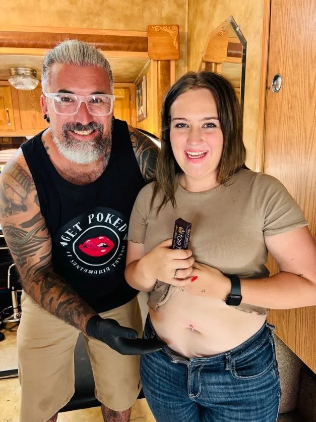 A tattooed man with gray hair, glasses, and a beard, smiling, standing next to a woman with brown hair and a beige shirt, who is holding a chocolate bar and has her shirt lifted to reveal her belly button piercing. They are indoors in a wood-paneled room.