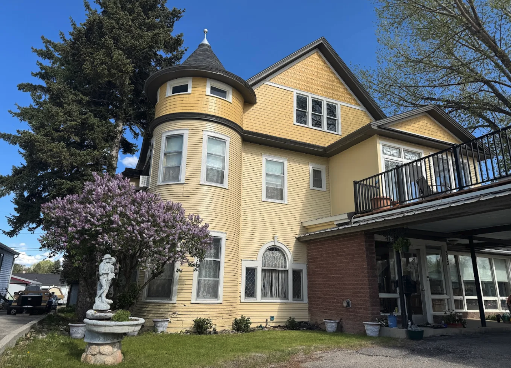 A large multi-story house with yellow and beige siding, a turret with a conical roof, several windows, some with lace curtains, a balcony, a flowering purple tree, a statue, and potted plants in the front yard.
