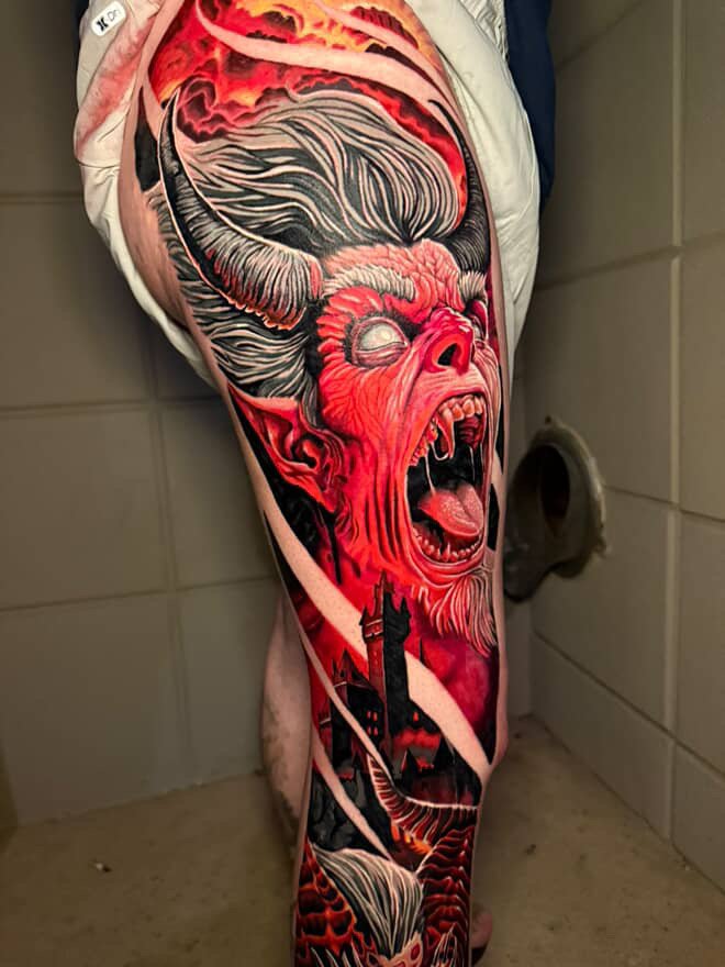 Colorful tattoo of a demonic figure with horns and an open mouth showing sharp teeth, set against a fiery red and orange background, covering part of a person's leg.