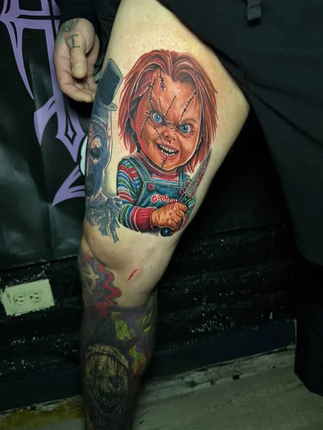 Colorful tattoo of a creepy doll with red hair, wearing striped sweater, holding a knife, with stitched scars on face and blue eyes.