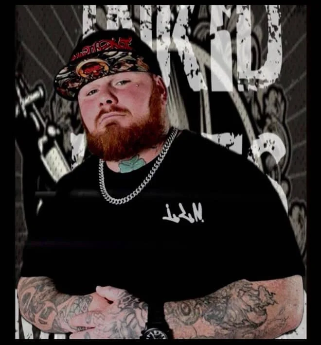A man with tattoos on his arms, a red beard, and a black cap with red and white designs. He is wearing a black T-shirt, a silver chain necklace, and has a tattooed neck. The background features graffiti-style art with large white letters and dark graphics.
