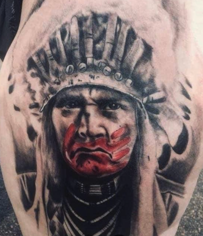 Tattoo of a Native American chief with a cherry red face paint, wearing a headdress made of feathers.