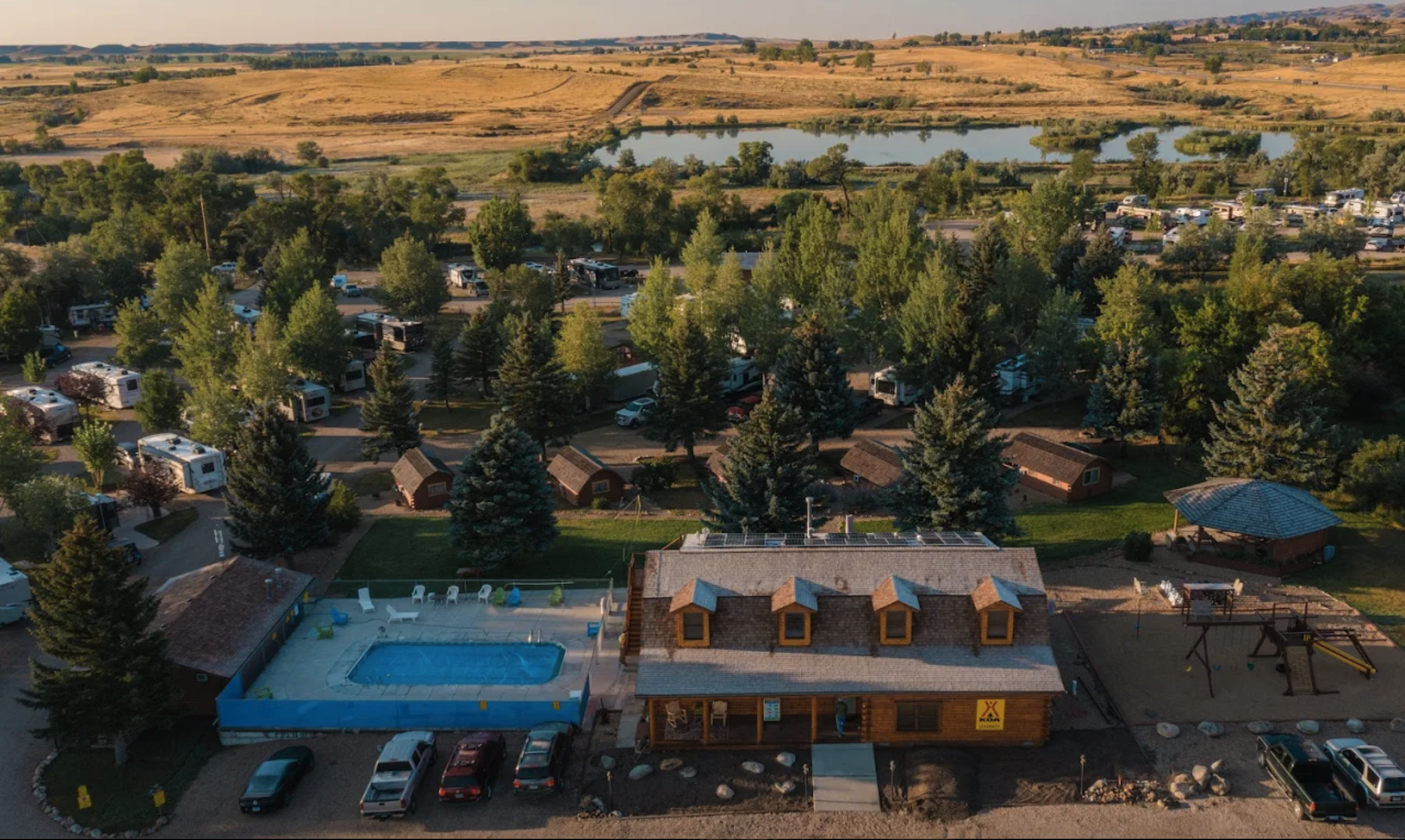 Aerial view of a campground with RVs and cabins, a swimming pool, a playground, and a building, surrounded by trees and open fields with a pond in the background.