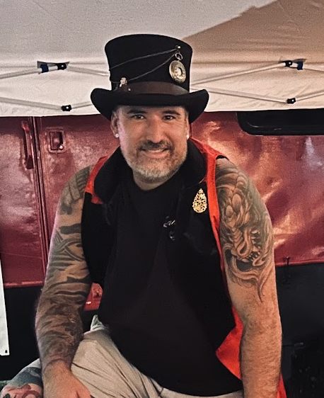 Man with a beard, wearing a black top hat with a badge, black vest with red trim, and displaying tattoos on both arms, sitting indoors in front of a red backdrop.