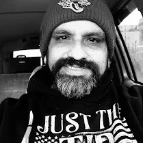 A man with a beard wearing a beanie hat and a hoodie, smiling inside a vehicle.