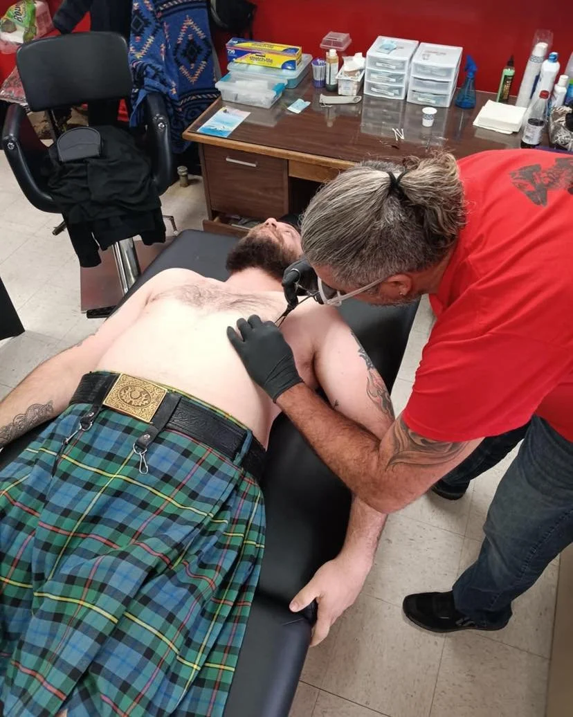 A man lying shirtless on an examination table while a healthcare professional, wearing glasses and gloves, uses a stethoscope on his chest in a medical office, with medical supplies and equipment on the desk nearby.