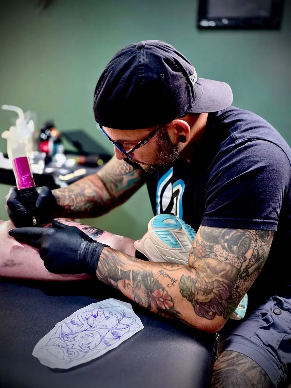 Tattoo artist in black cap and gloves tattooing a person's arm on a black surface with tattoo equipment in the background.