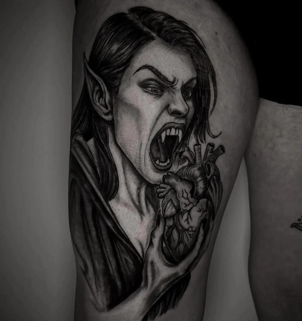 A black and gray tattoo of a fierce woman with pointed ears, long flowing hair, and a snarling expression. She appears to be screaming or shouting, with her mouth wide open revealing sharp teeth. There is an anatomical heart tattoo below her face, with detailed shading.
