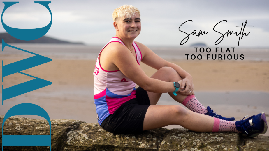 A person with short blonde hair sitting on a rock at the beach, wearing a pink, white, and blue sports top, black shorts, colorful socks, and sneakers, smiling at the camera with overcast skies in the background, next to the text 'Sam Smith' and 'Too Flat Too Furious'. The name 'DOVE' is partially visible on the left side of the image.