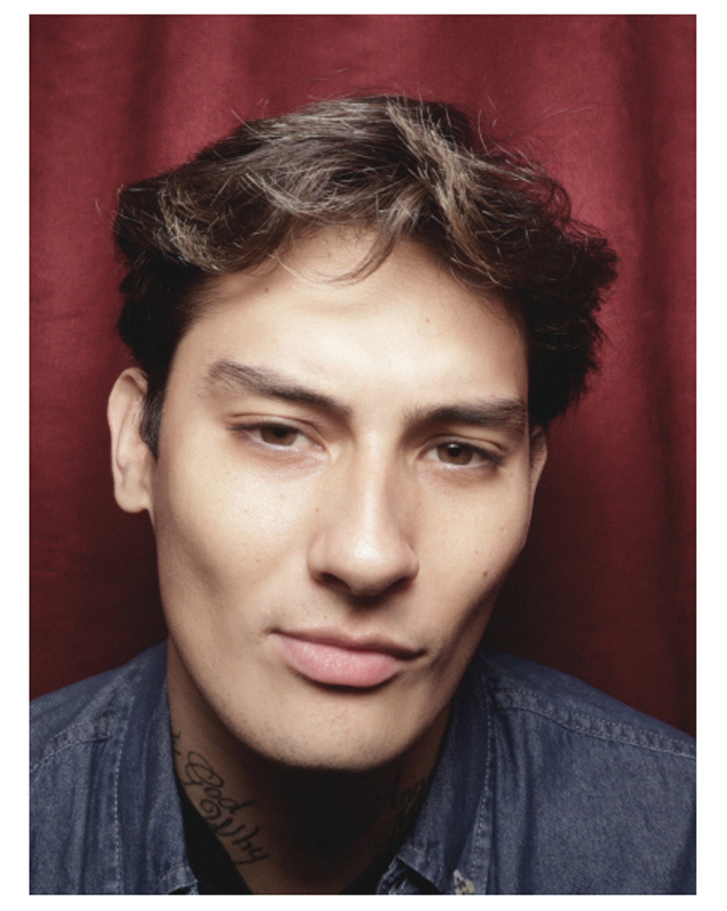 Portrait of a person with brown hair wearing a denim shirt against a red backdrop.