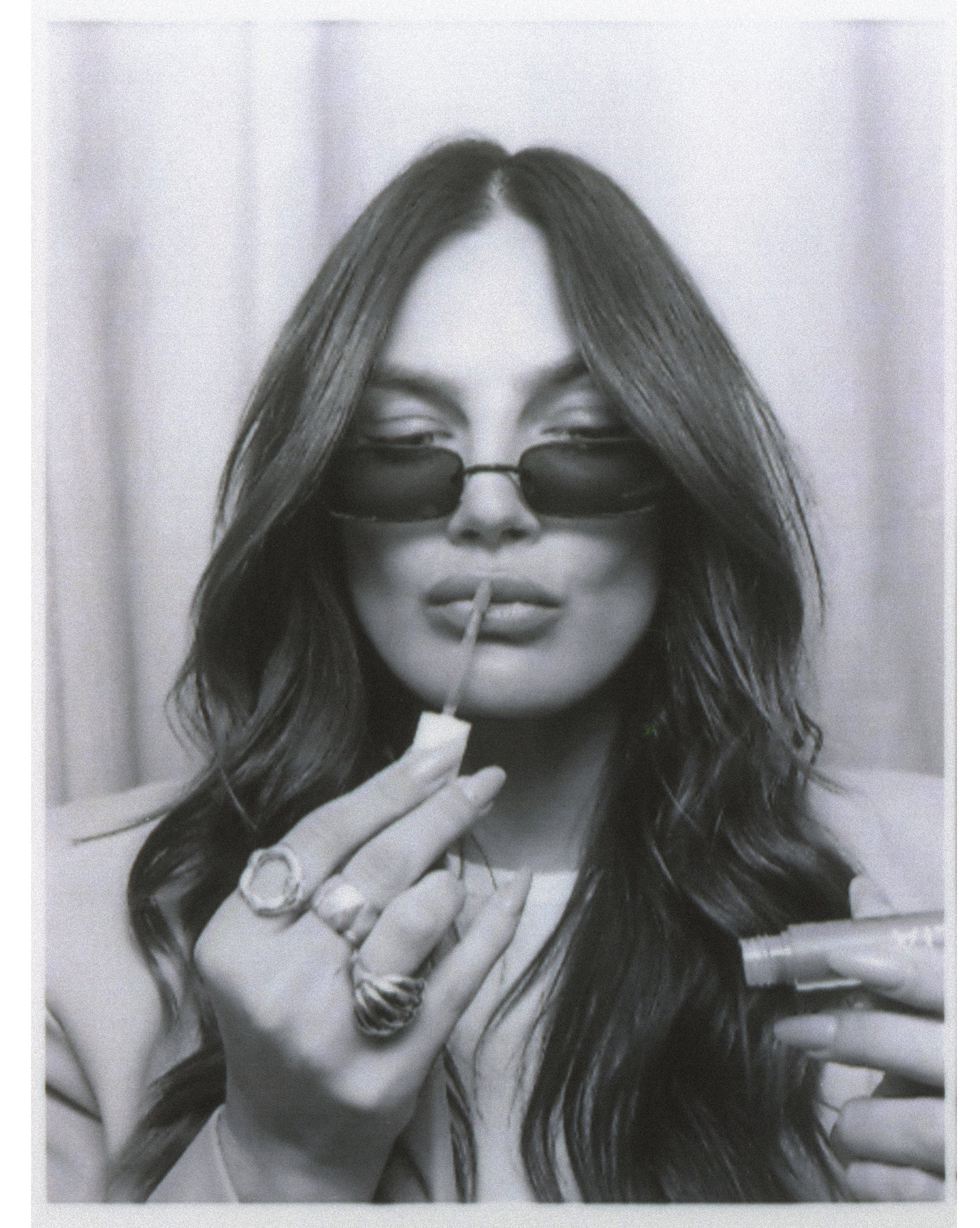 Woman with long hair wearing sunglasses applying lip gloss, holding applicator and tube, black and white photo.