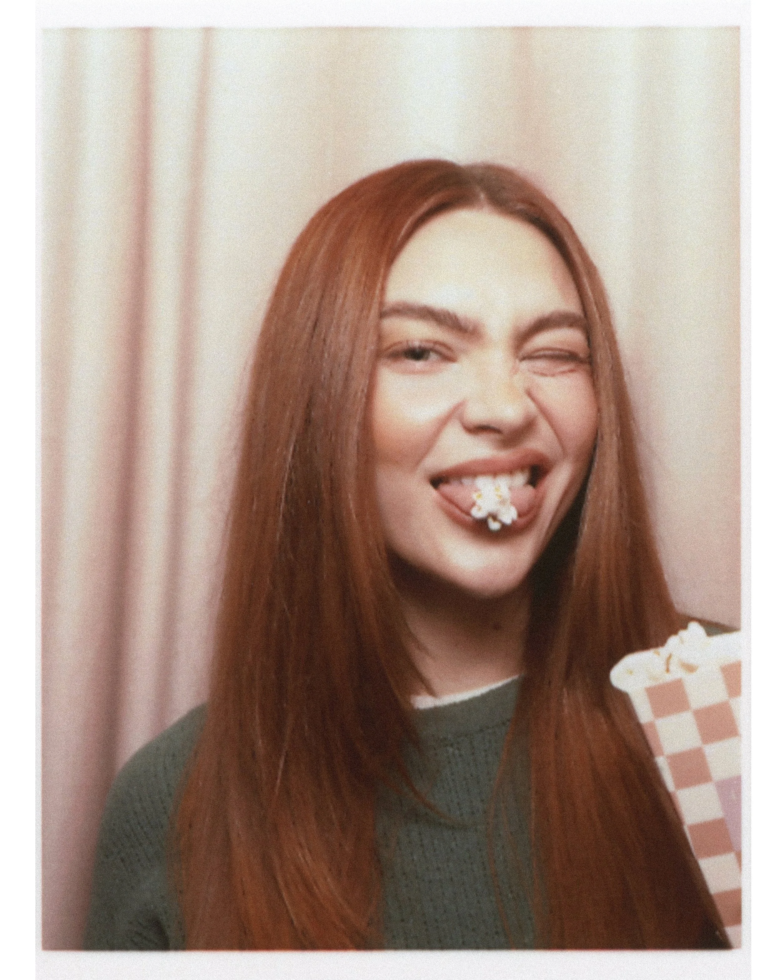 Person with red hair holding popcorn, winking with popcorn in mouth, wearing green sweater.