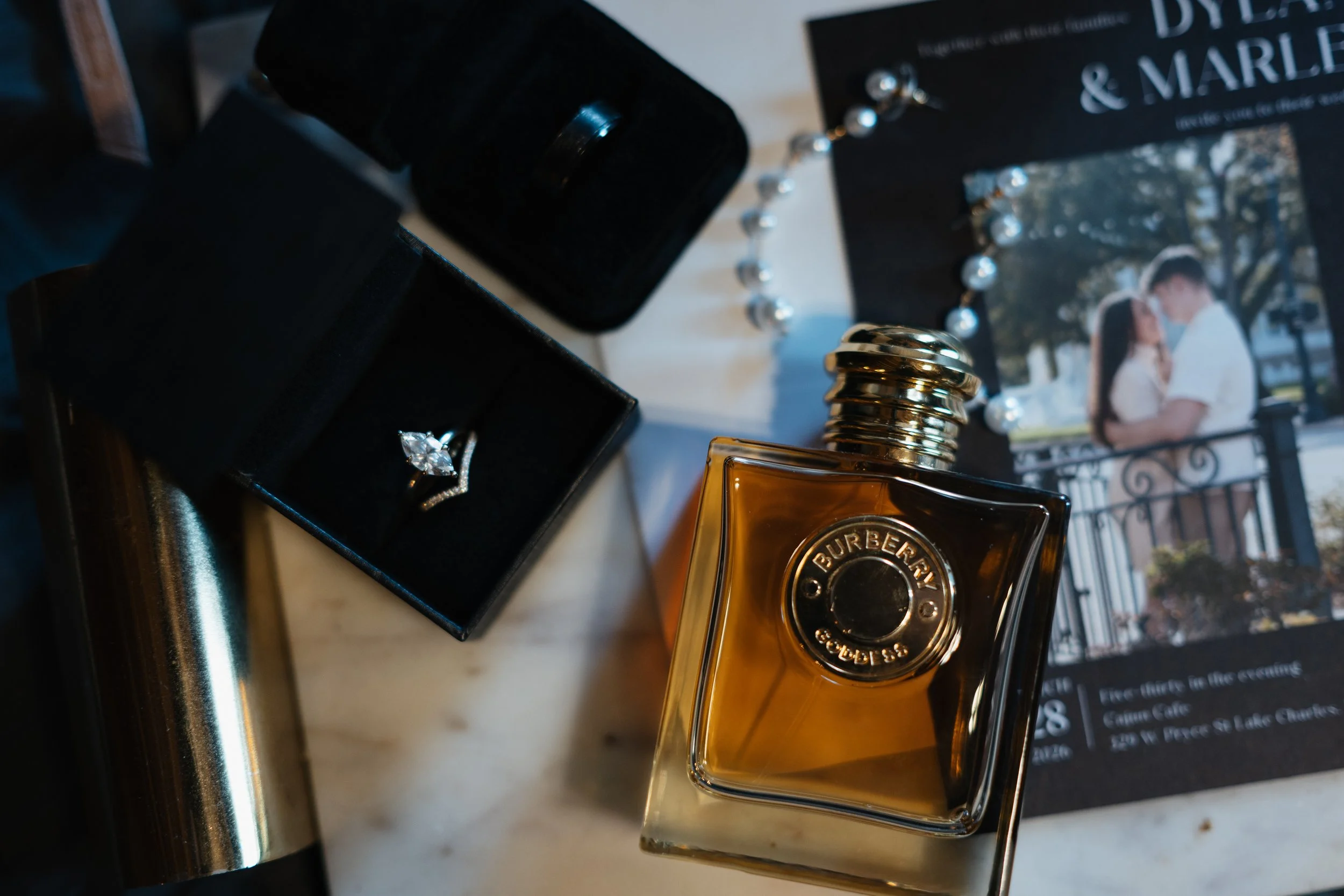 A bottle of Burberry perfume, a black jewelry box with a diamond ring, and a wedding invitation with a photo of a couple.