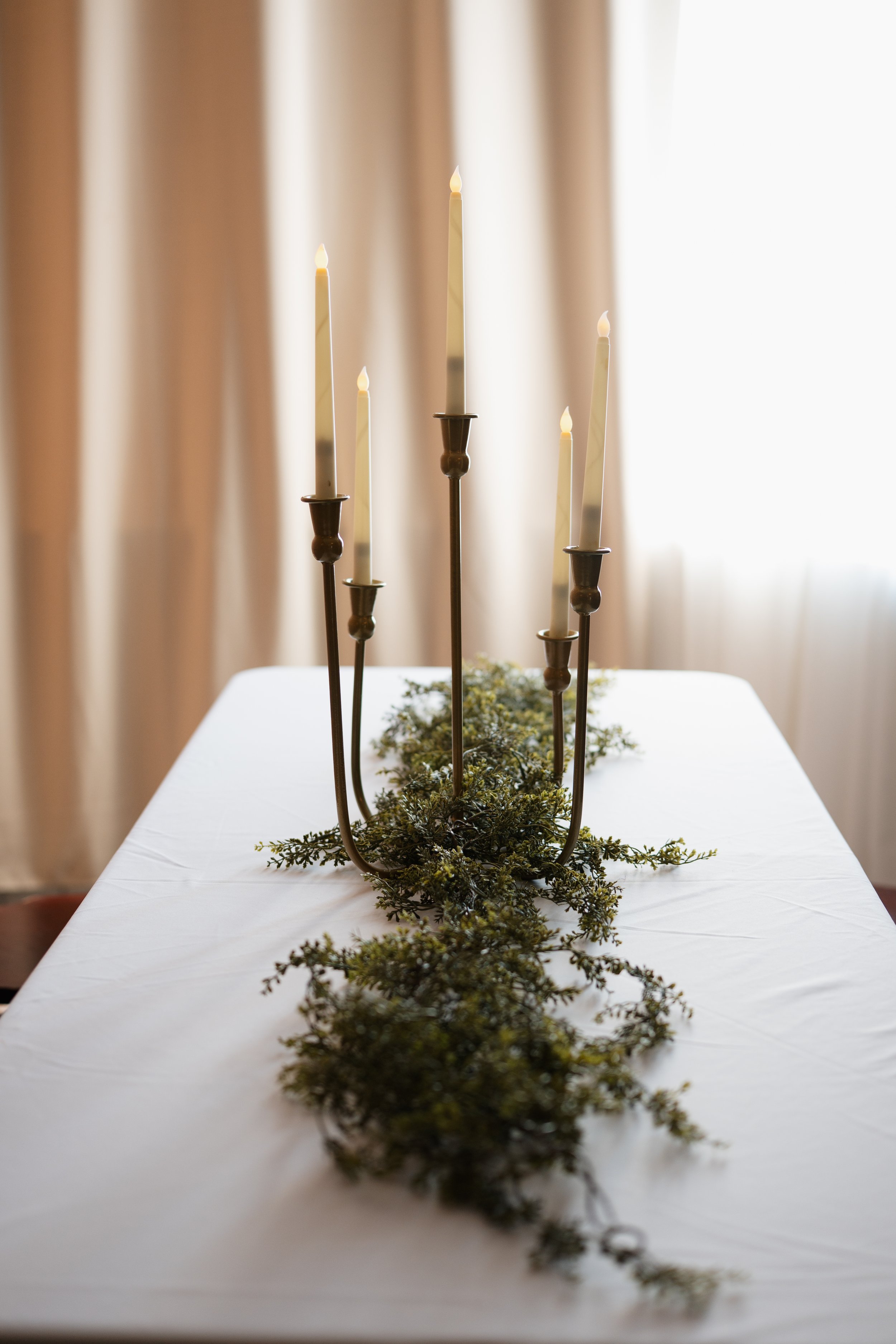 A table centerpiece with five tall candlesticks, some with lit candles, and greenery on a white tablecloth.
