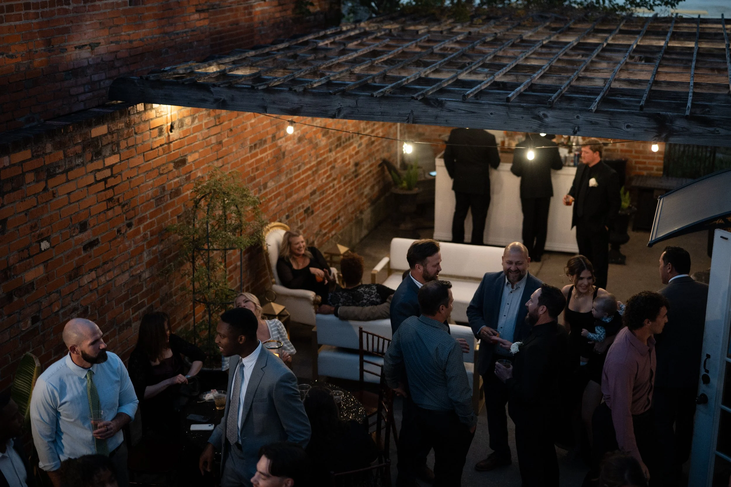 Photograph of people socializing at an outdoor event in an enclosed patio with string lights, brick walls, and a white sofa at the Cajun Cafe in Lake Charles, Louisiana.