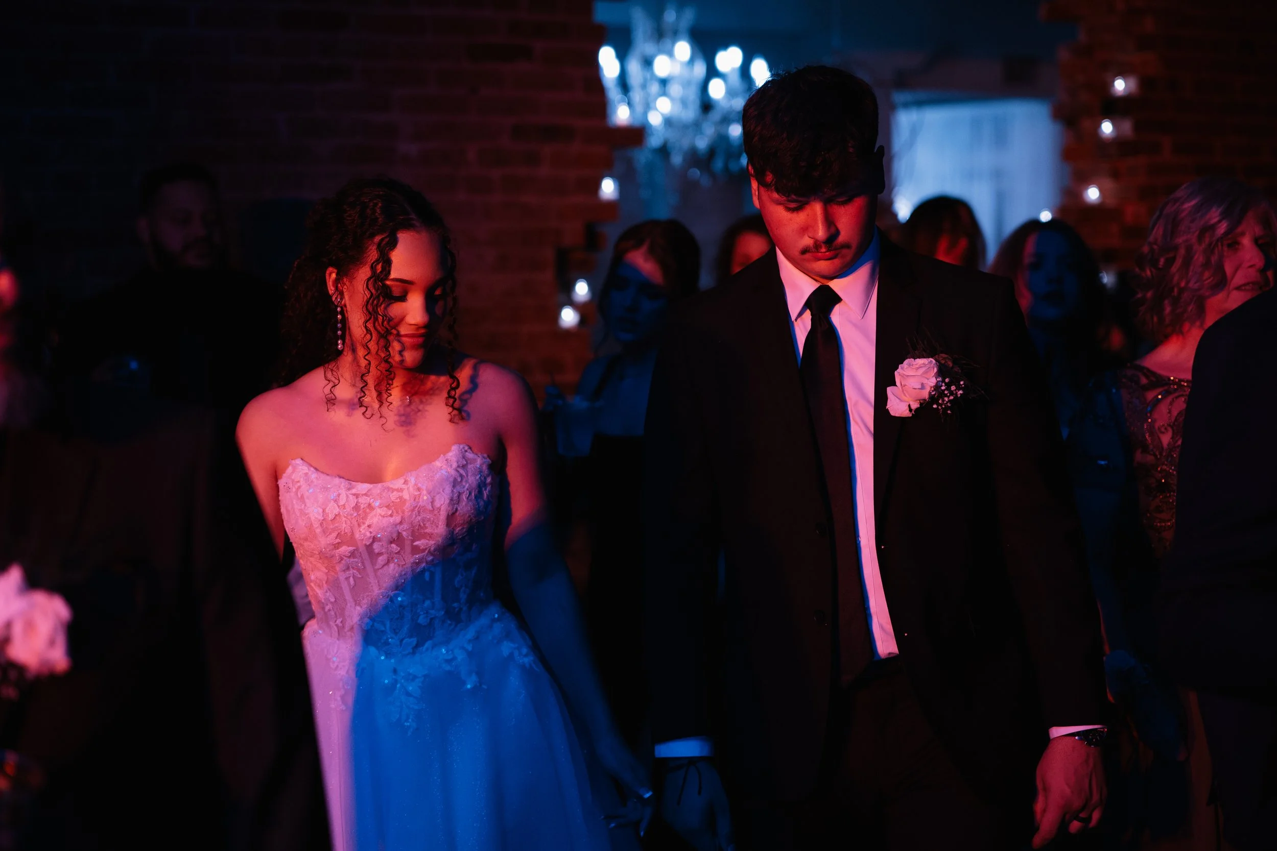 A young woman in a white wedding dress and a young man in a black suit with a white shirt and black tie, both with pink flowers on their lapels, standing close together in a dimly lit room with other people and a chandelier in the background.