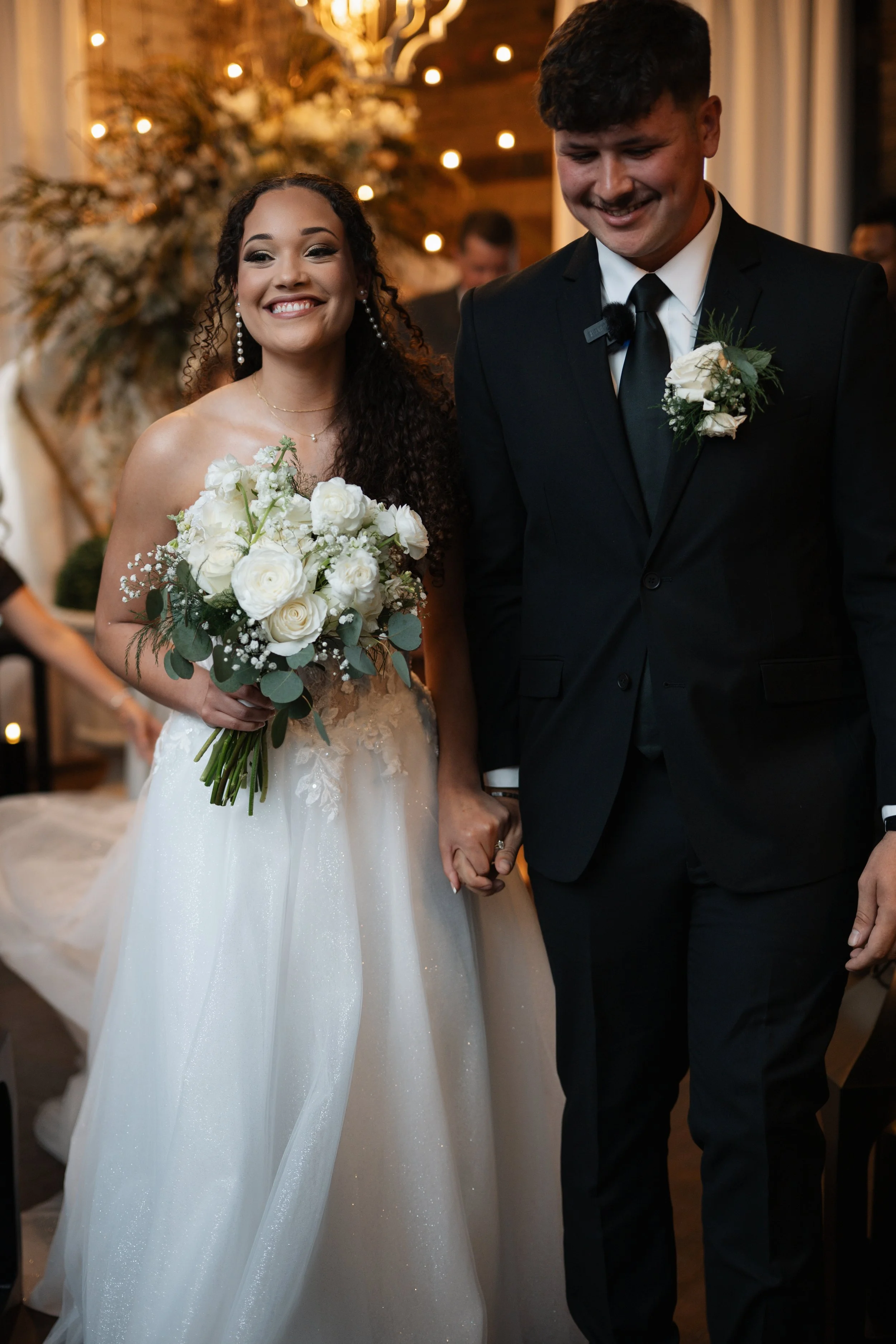 Bride and groom holding hands during a wedding ceremony; bride in a white wedding gown holding a bouquet of white flowers, groom in a black suit with boutonniere; warm, decorated indoor setting.