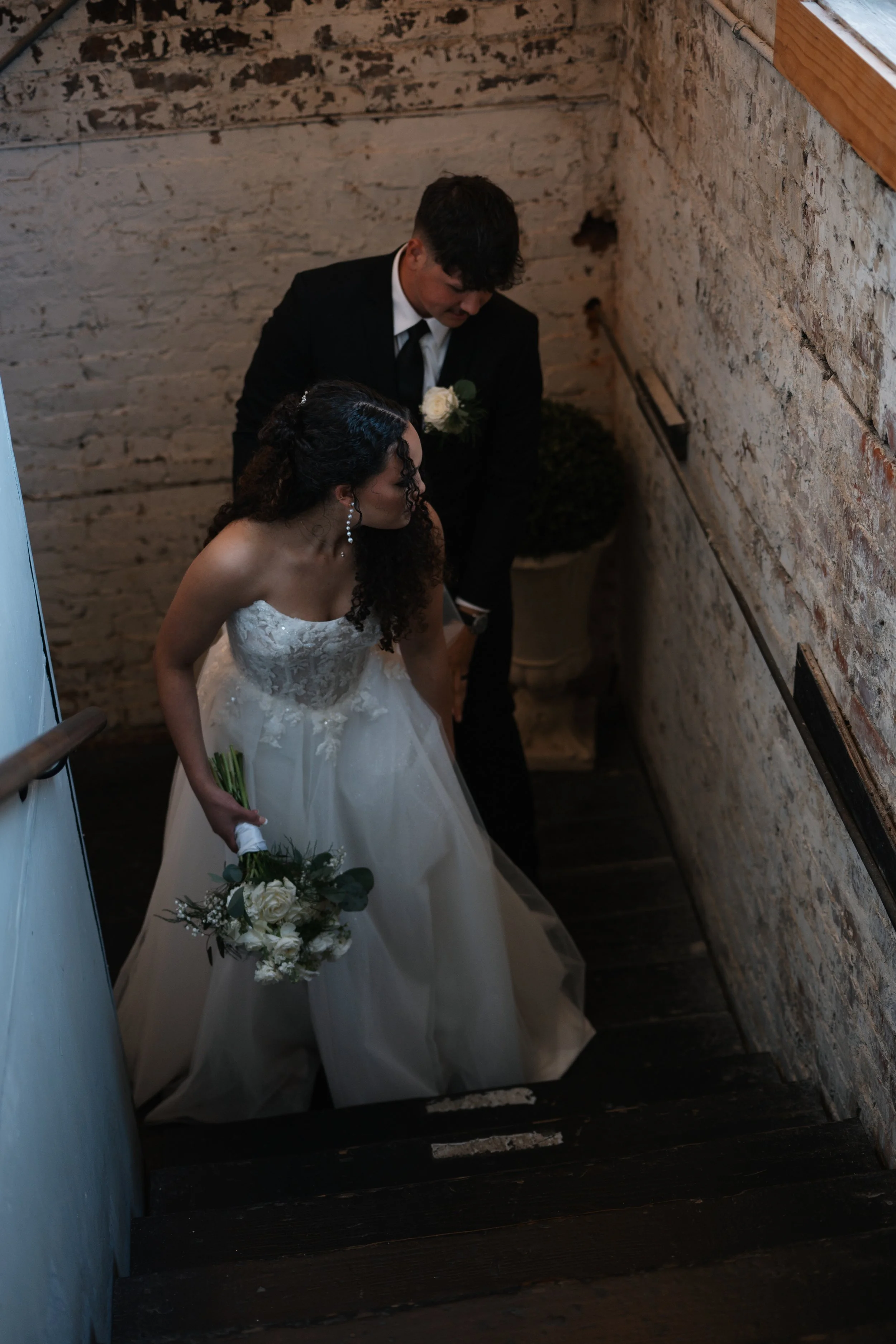 A bride and groom descend a dark staircase, with the bride holding a bouquet of white flowers. The bride is wearing a white strapless wedding gown with lace details, and the groom is dressed in a black suit with a white boutonnière. They are in an in