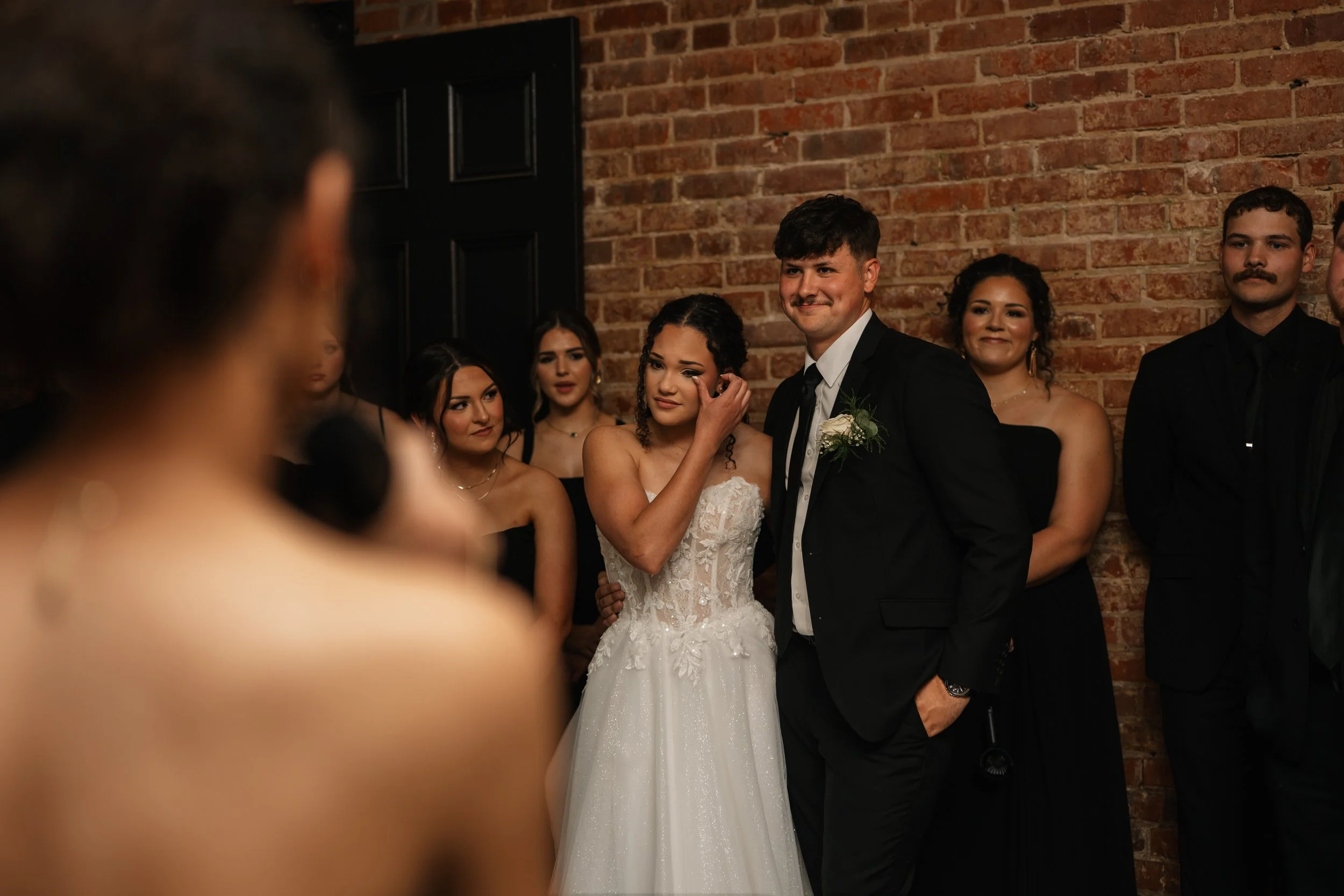 Bride and groom standing together during wedding ceremony, surrounded by bridesmaids and groomsmen, with a brick wall in the background.