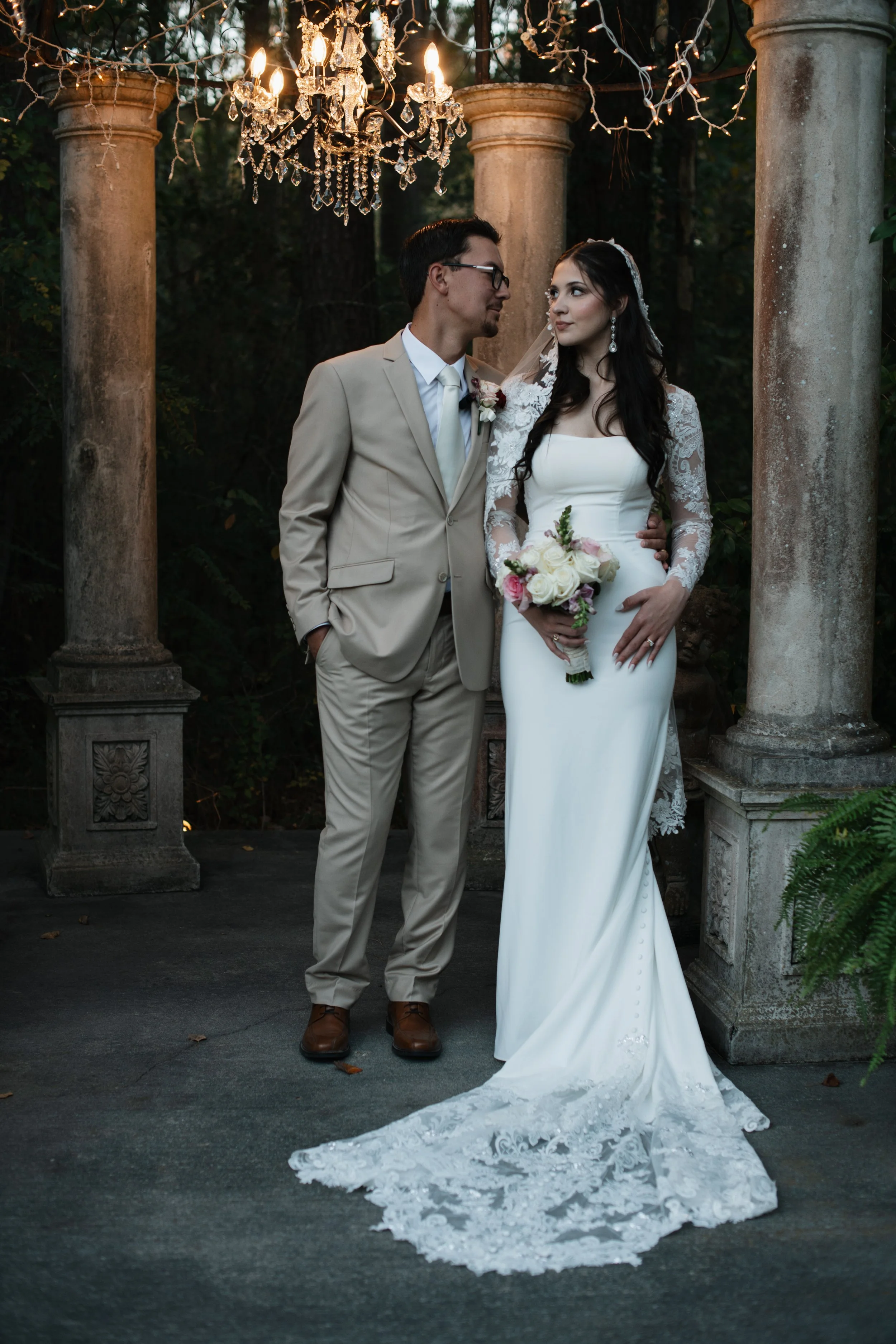 A bride and groom standing together outdoors under a chandelier, with columns and greenery in the background, dressed in wedding attire.