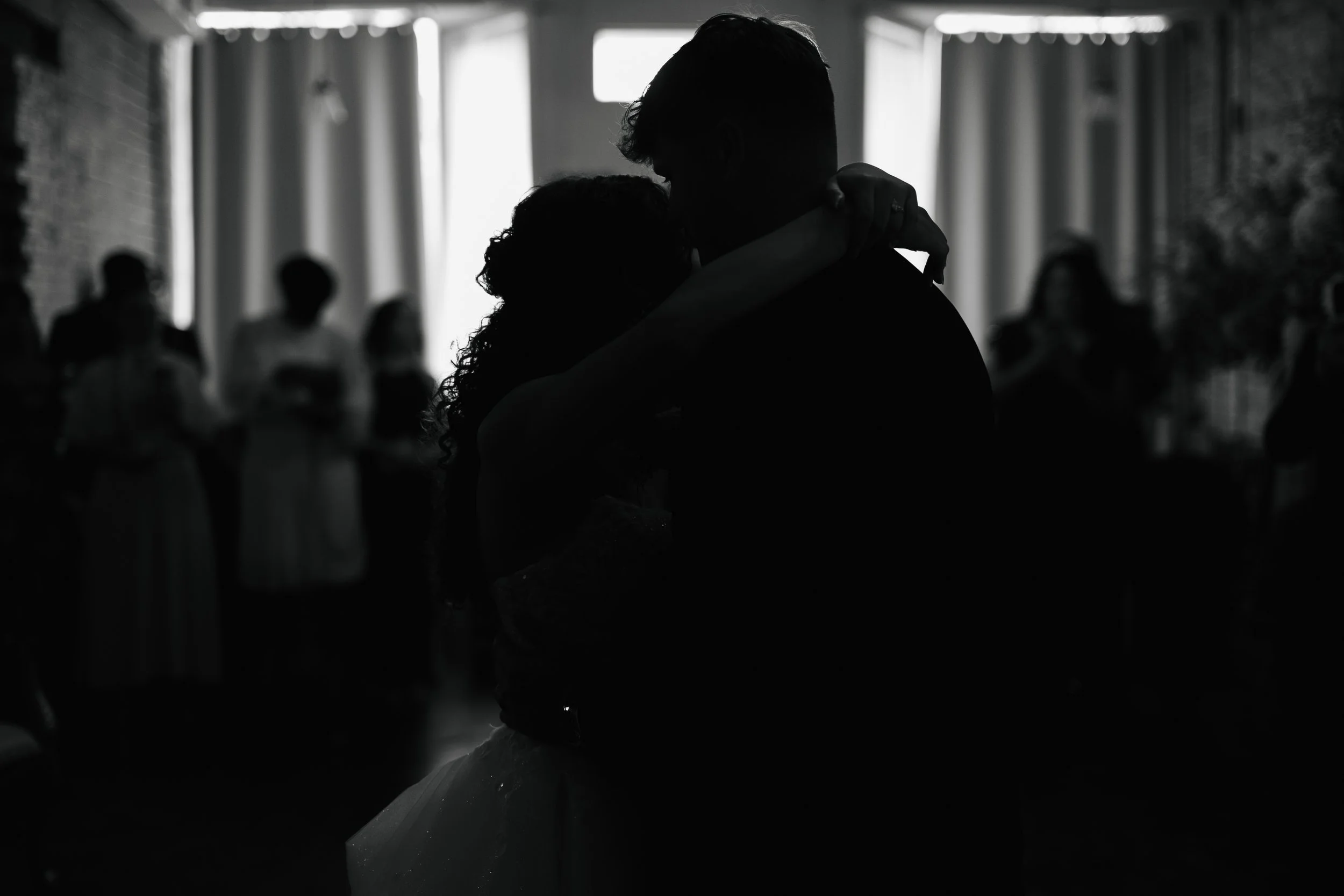 Silhouette of a couple dancing closely at their wedding reception, with guests in the background.