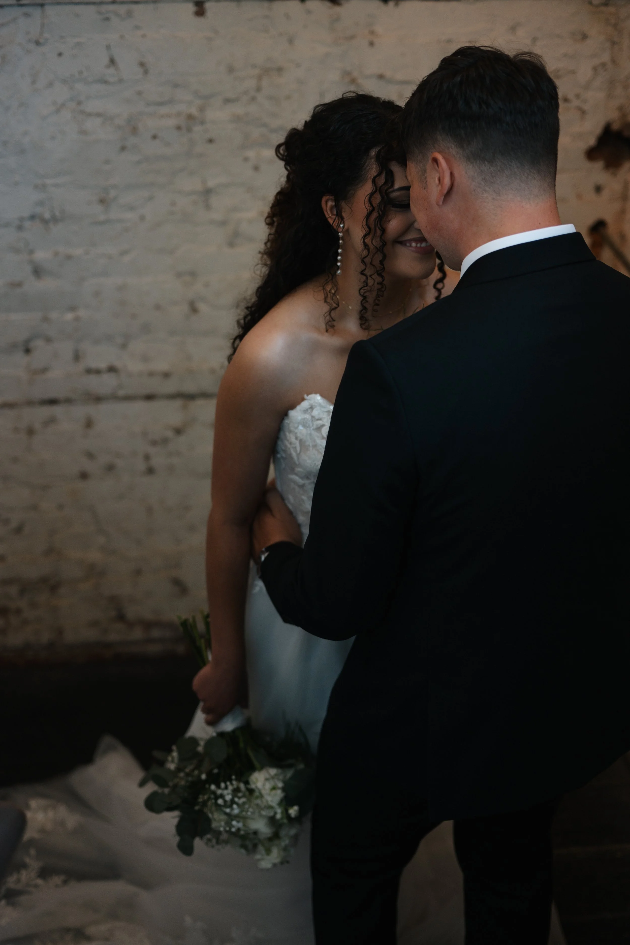 Portrait of a bride and groom standing close together, facing each other with foreheads touching, after their wedding ceremony. The bride is holding a bouquet of flowers. The background features a textured brick wall.