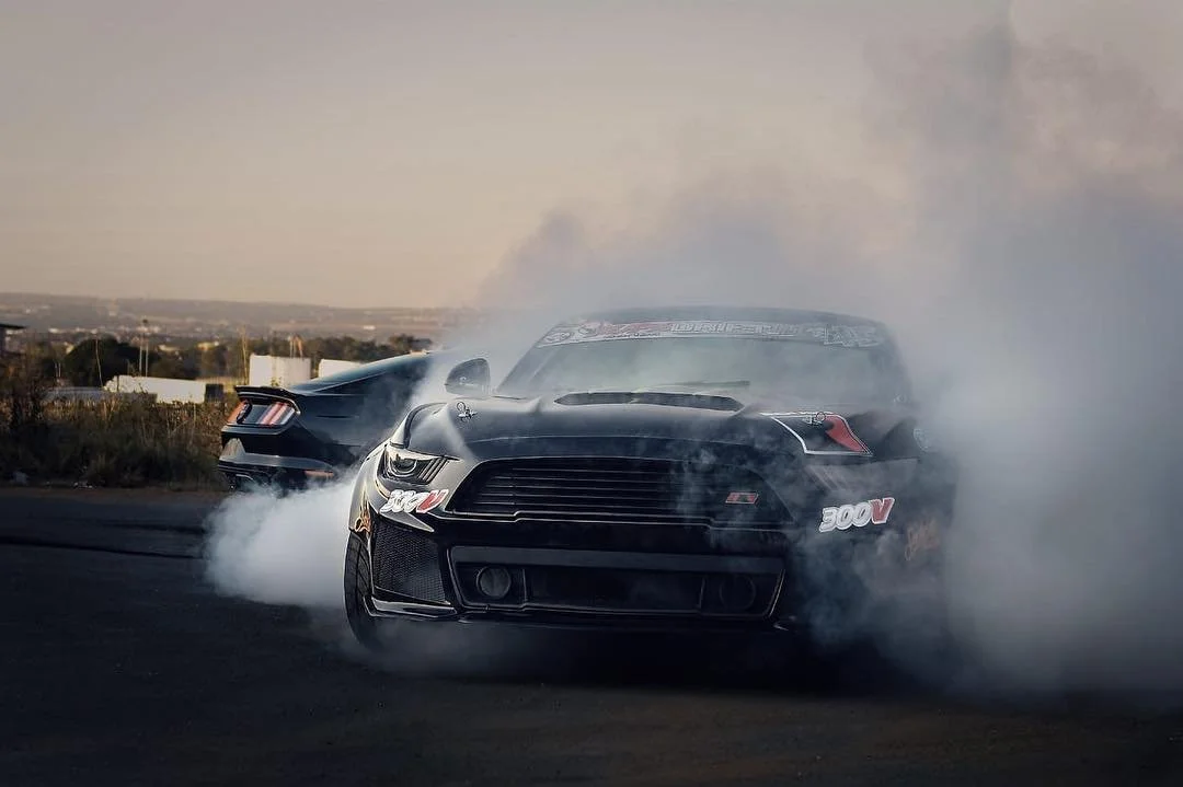 Two black sports cars performing burnouts on a paved road, with smoke and tire smoke filling the air and a distant landscape in the background.
