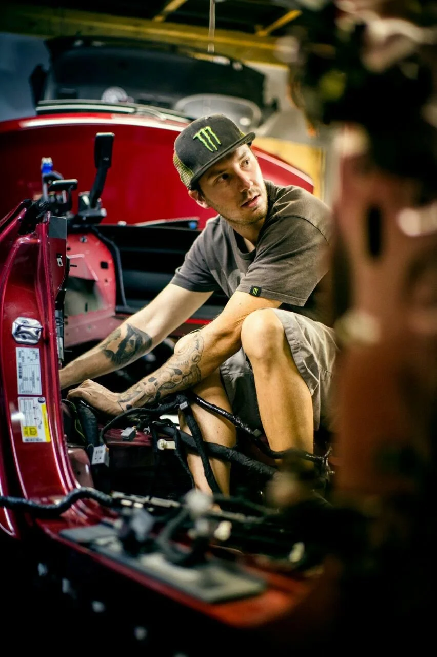 A young man with tattoos wearing a black Monster Energy cap and gray T-shirt working on the engine of a red race car inside a garage, with another person blurred in the foreground.