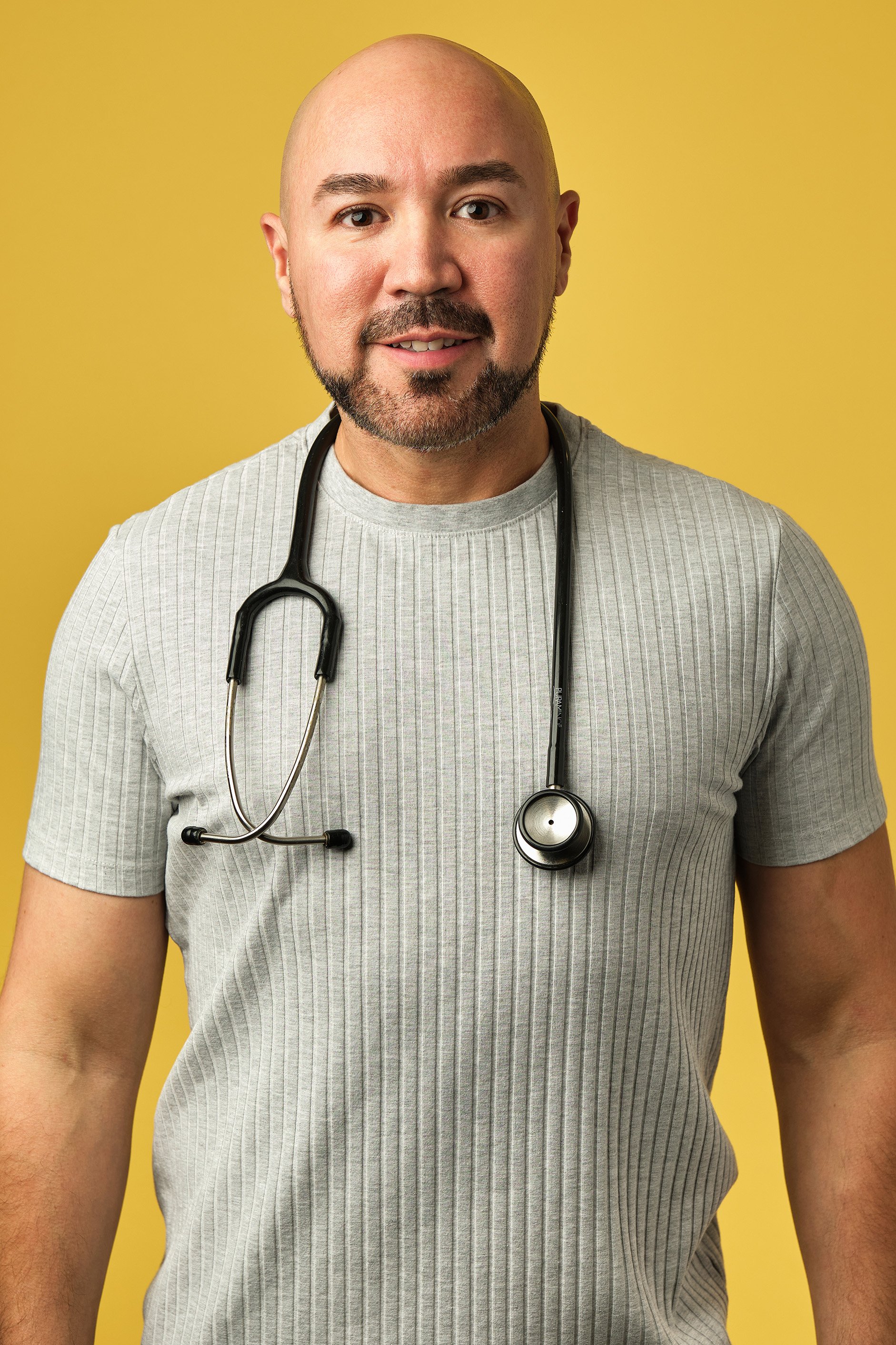 Hombre calvo con barba, vistiendo camiseta gris, con estetoscopio alrededor del cuello, fondo amarillo.