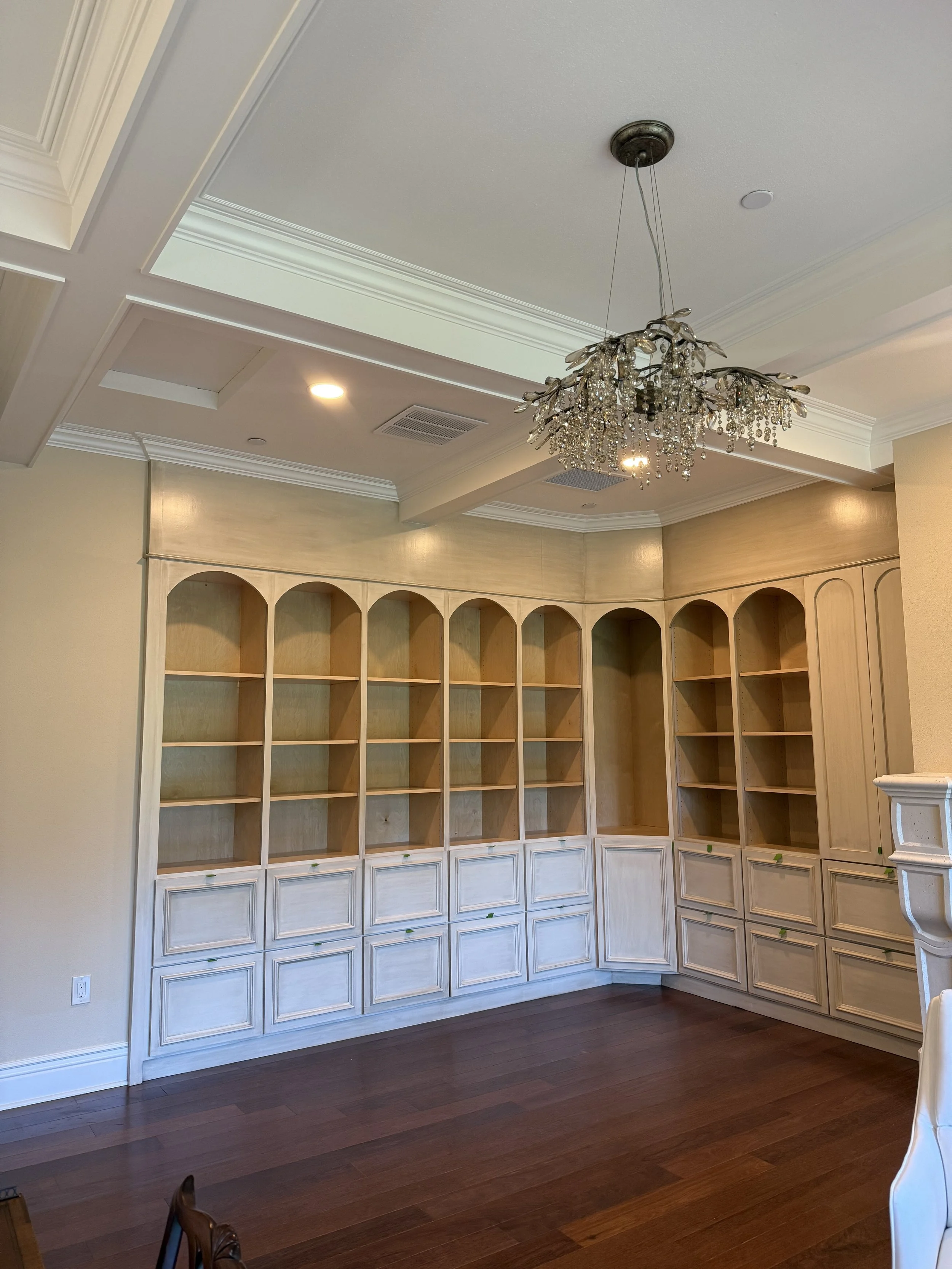Empty built-in wooden bookshelf with arched top compartments in a room with cream-colored walls, a chandelier hanging from the ceiling, and hardwood flooring.